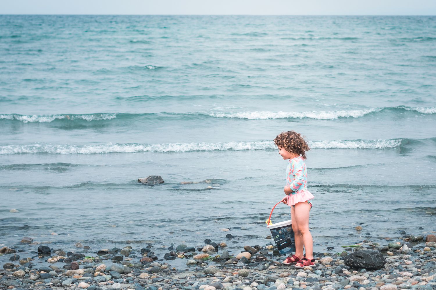 A toddler girl stands on the rocky beach with her bucket as waves from the ocean appear behind her. Parksville, BC.