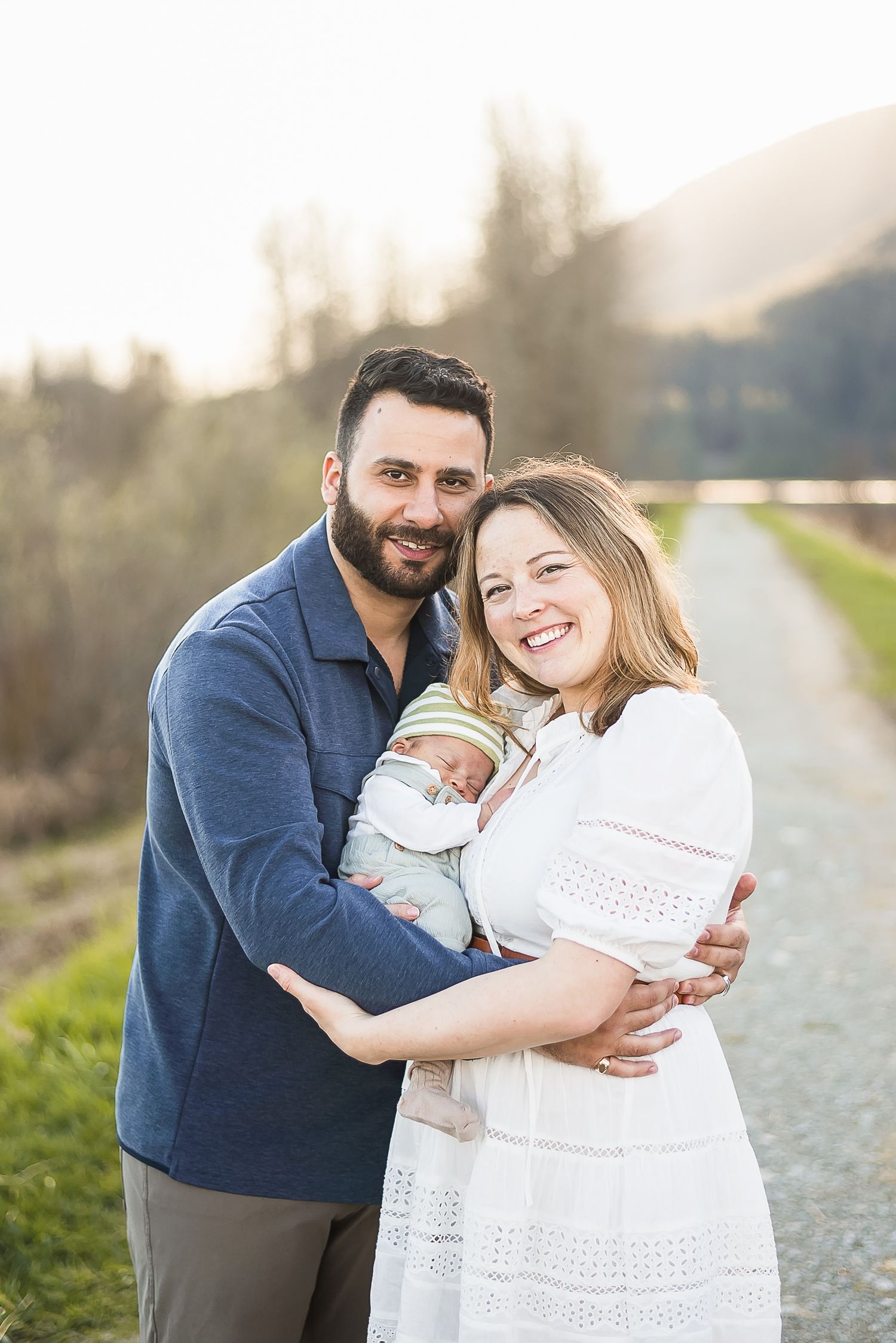 A couple embrace their 3 week old baby in the mountains on a sunny spring afternoon, golden light shining behind them.
