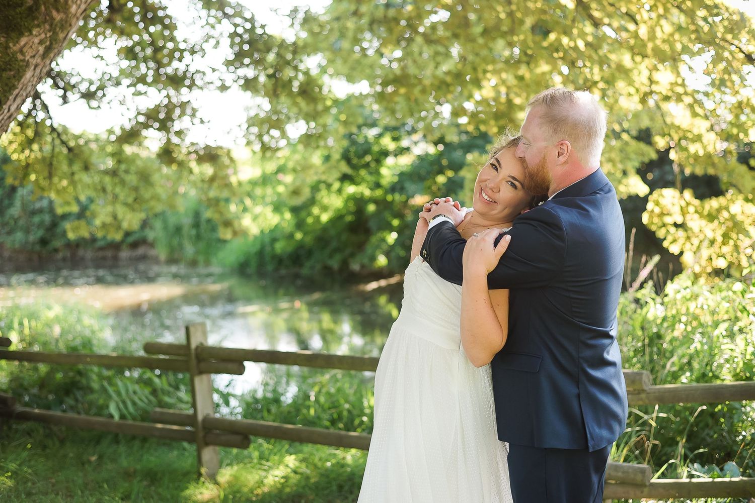 In the shade of the trees, on a July afternoon, a groom wraps his arms around his bride, while she smiles at the camera.