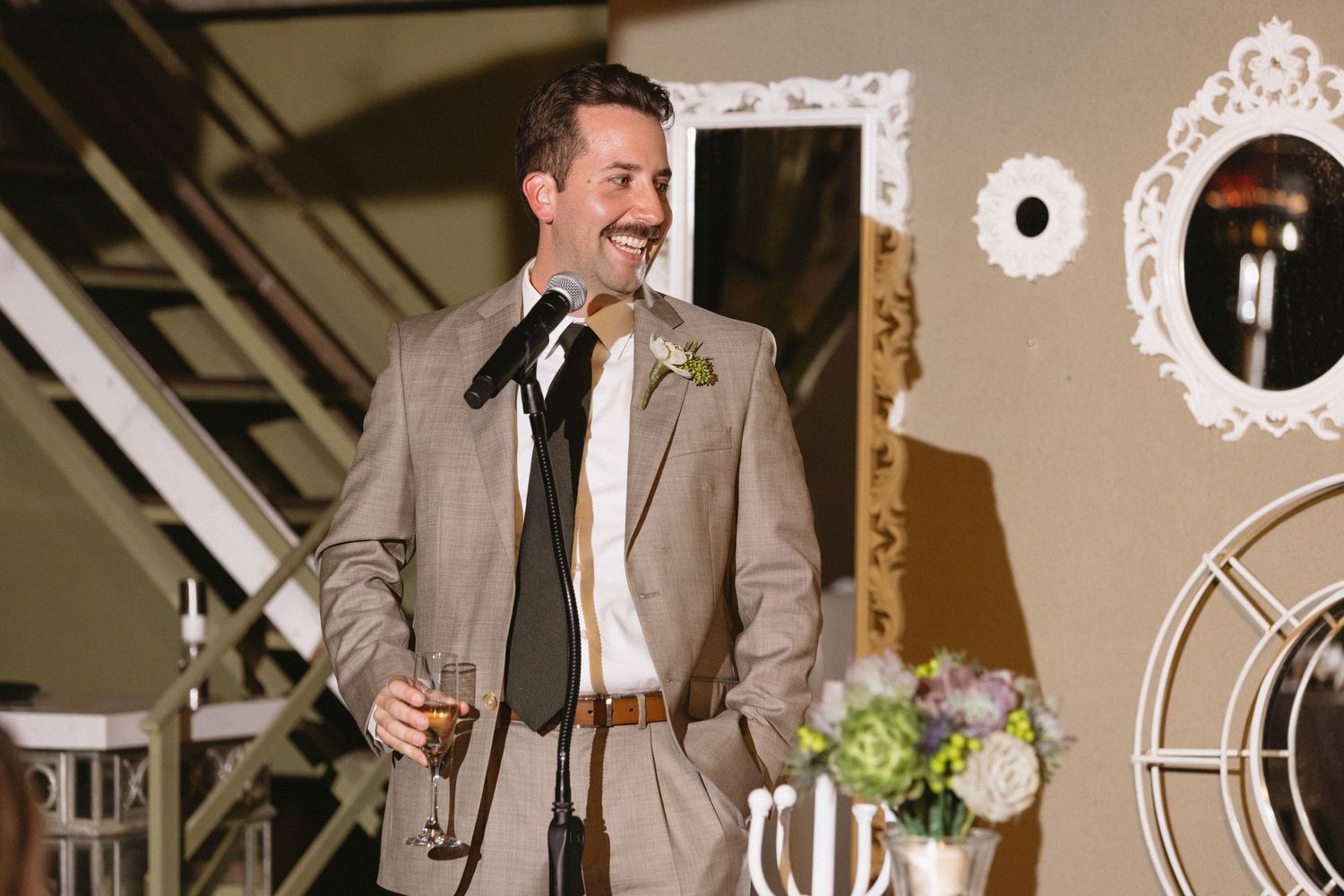 Smiling groom in gray suit with boutonniere holds a drink while giving a speech at an elegant wedding reception.