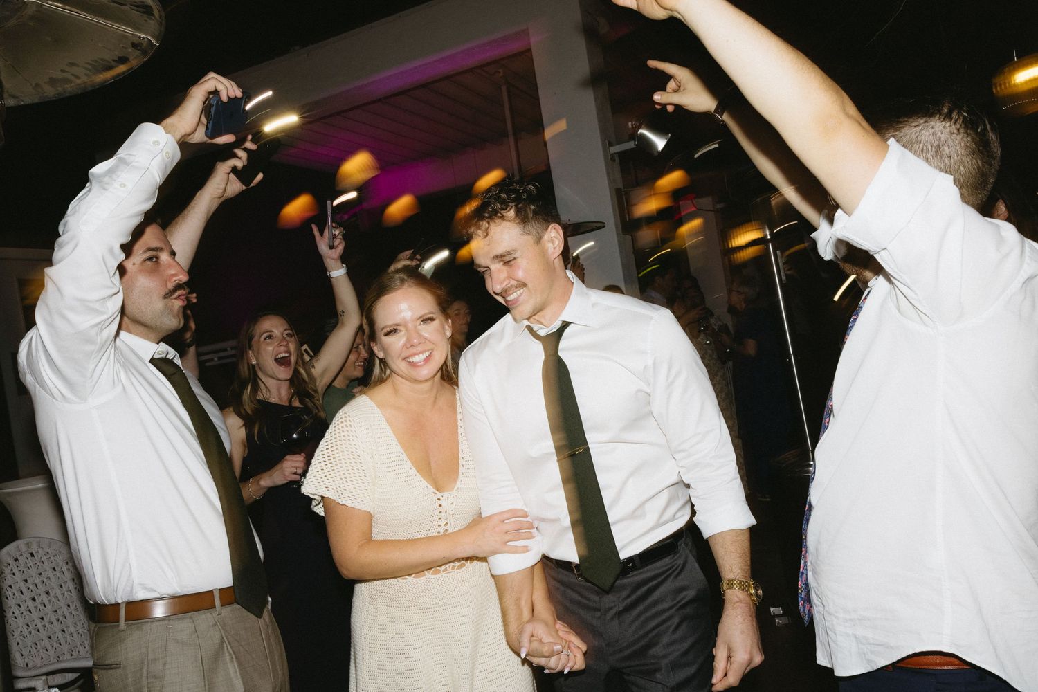 Bride and groom dancing joyfully at wedding reception surrounded by celebrating guests raising hands.