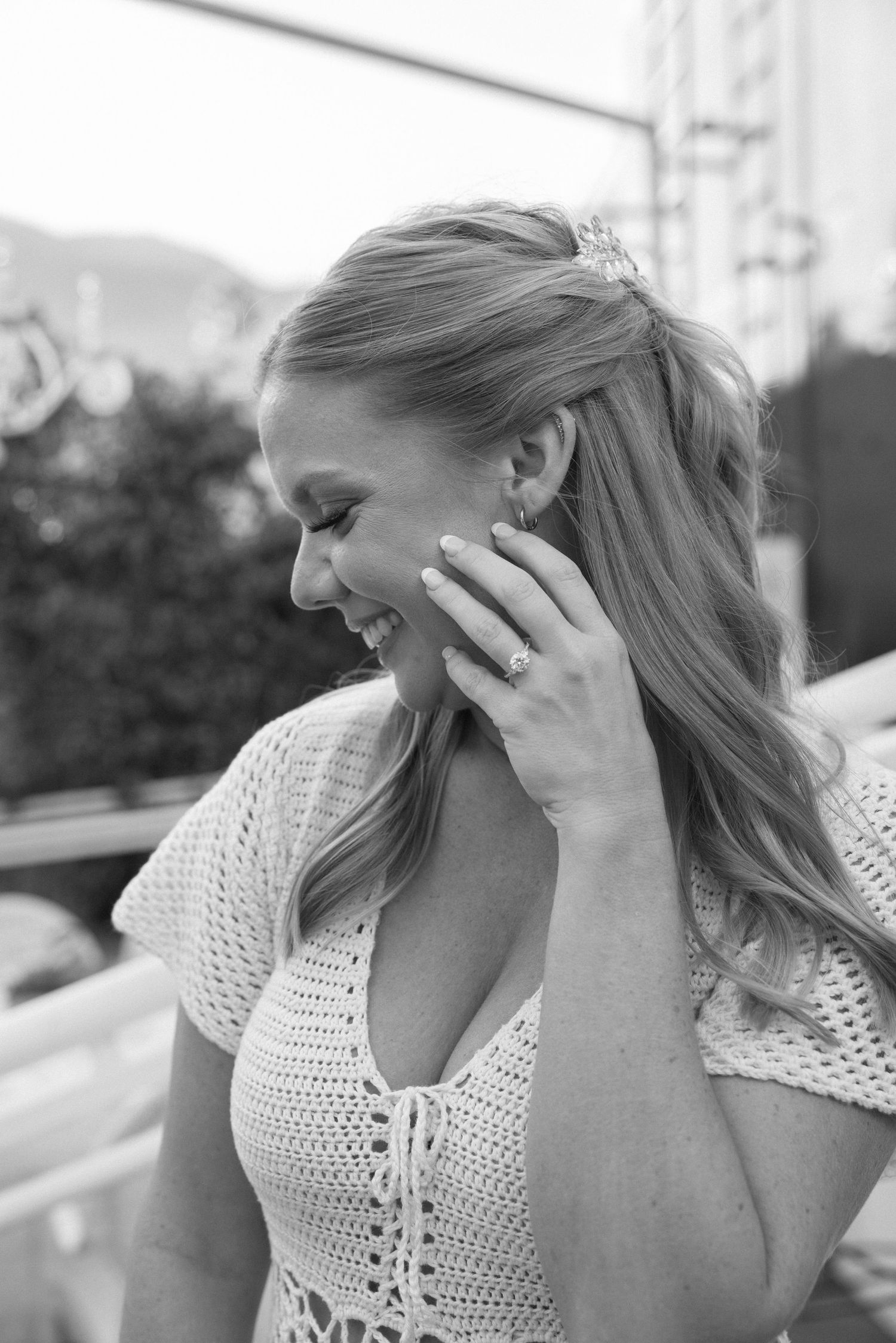 Black and white photo of a smiling blonde woman in a crochet top, touching her face, wearing a ring and earrings.