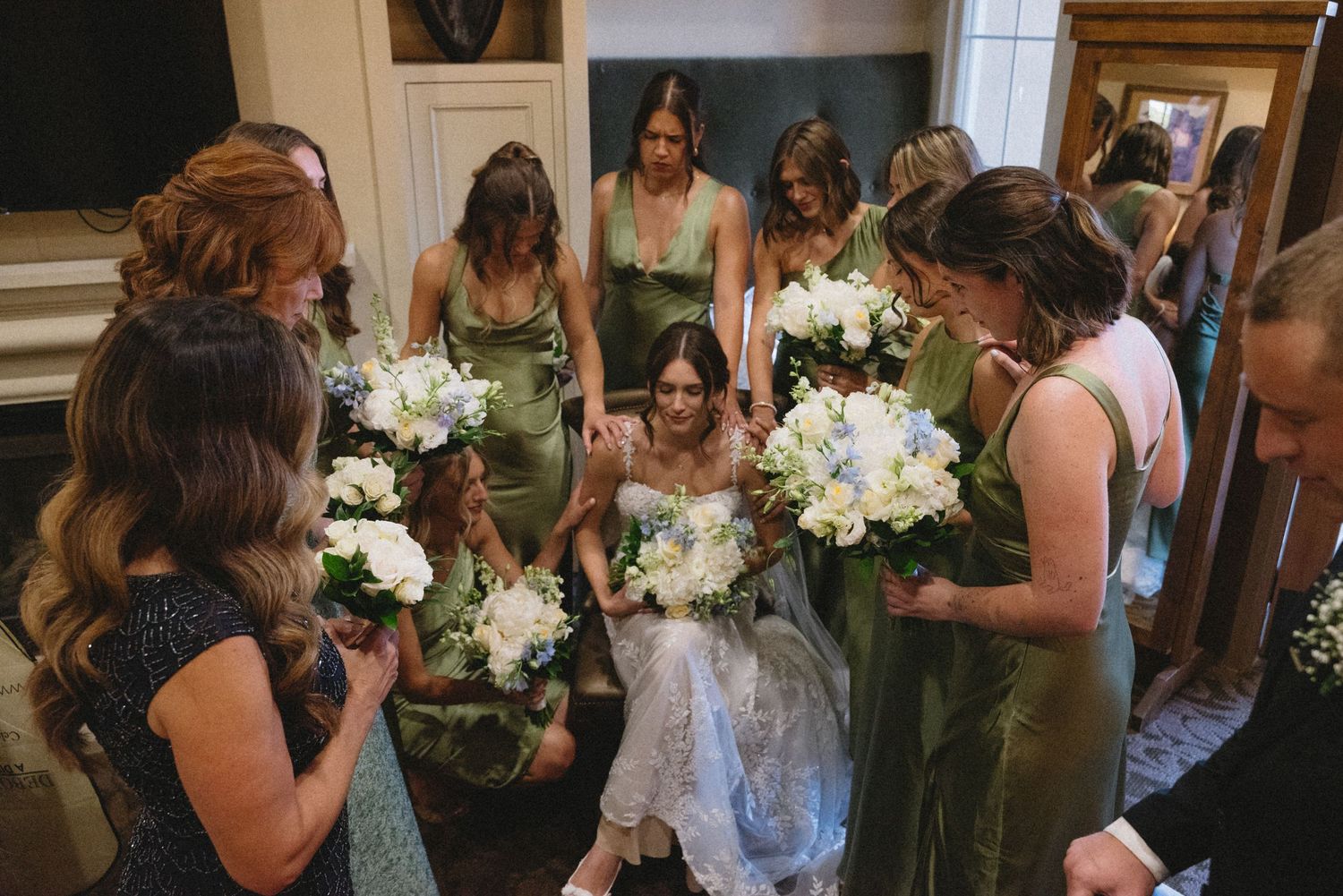 Bride surrounded by bridesmaids in sage green dresses holding white and blue bouquets in an elegant indoor setting.