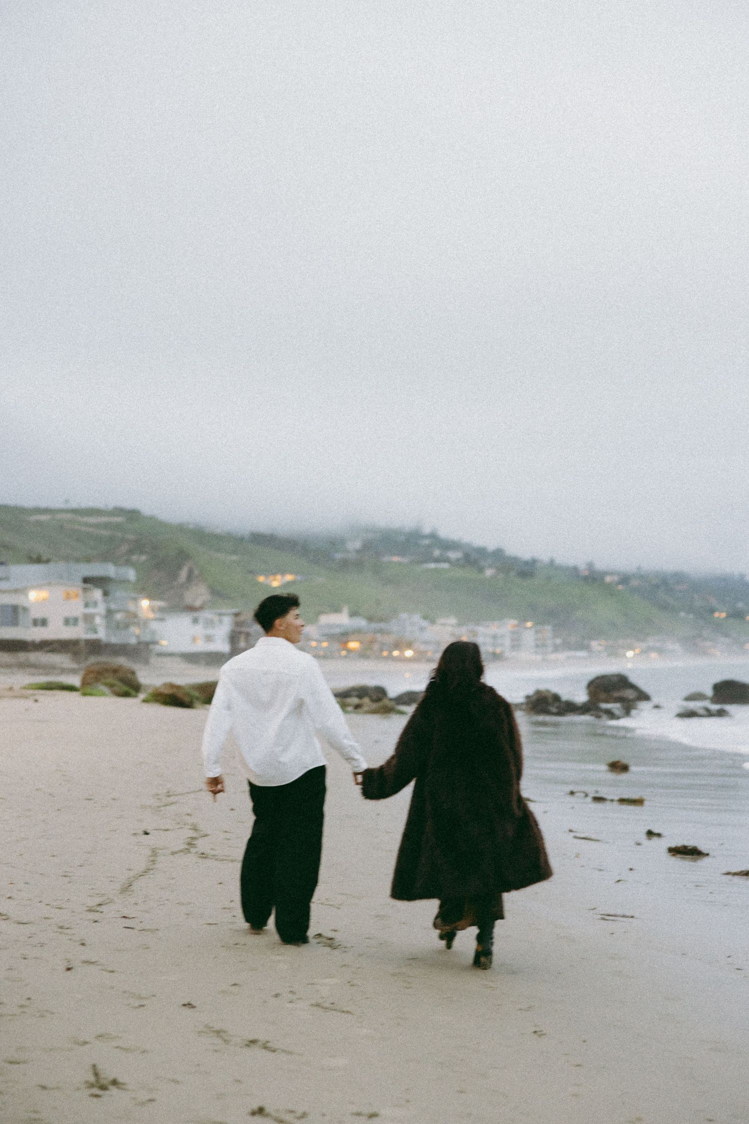 Couple in dark clothing walking together on foggy beach.