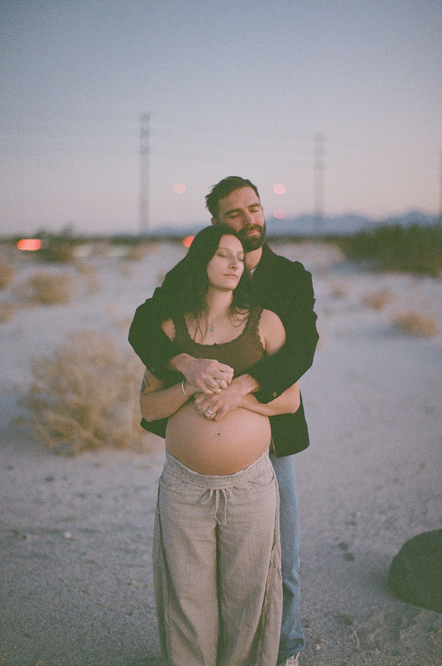 A film maternity photoshoot in a desert landscape with a couple embracing at sunset.