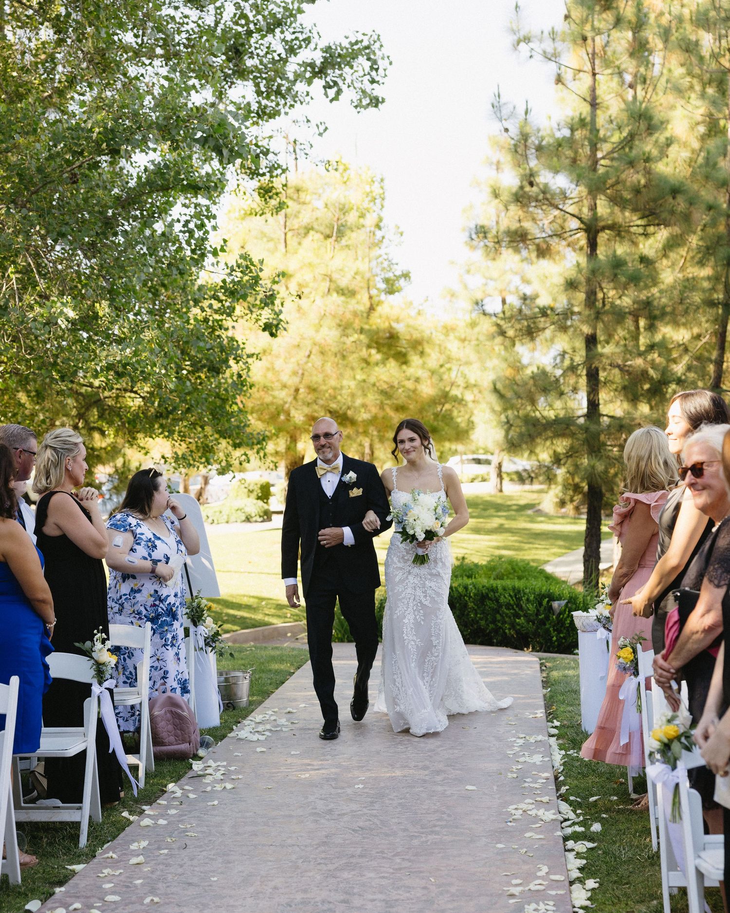 Bride walks down outdoor aisle with escort at sunny garden wedding ceremony, guests seated on white chairs among lush trees.