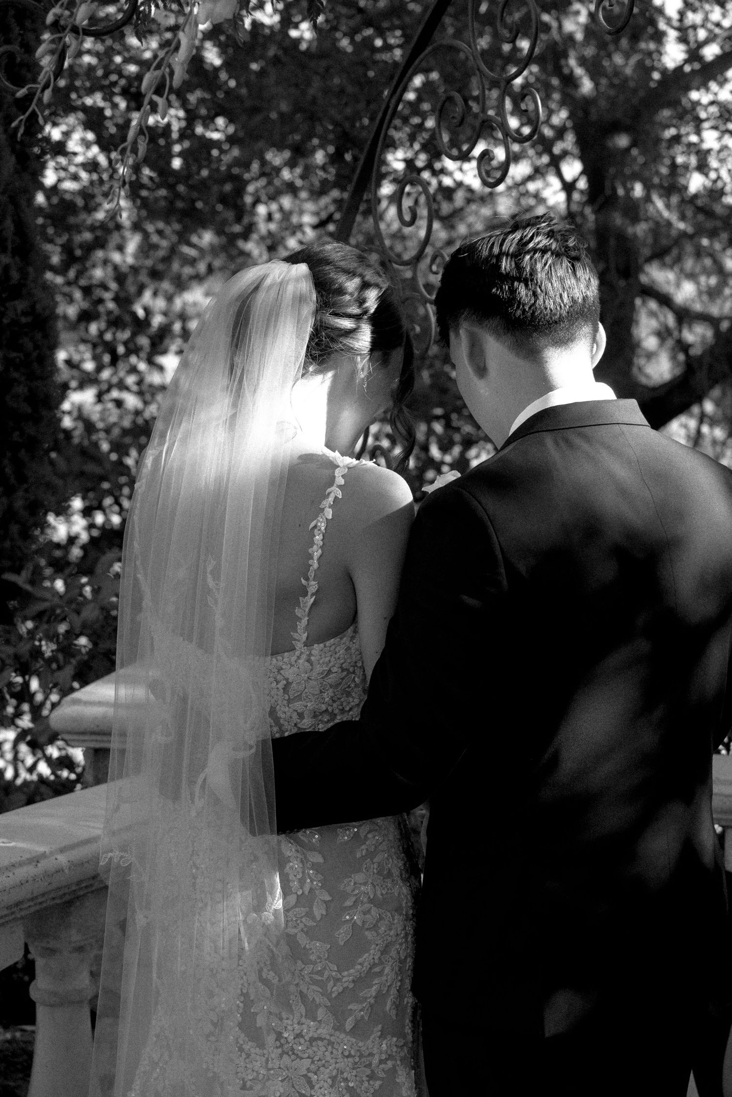Black and white photo of bride in lace gown and veil standing with groom in tuxedo at outdoor wedding ceremony.