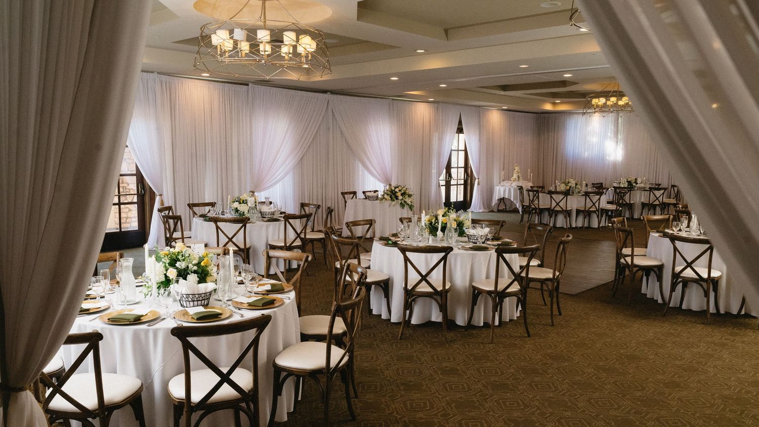 Elegant wedding reception hall with round tables, white linens, floral centerpieces, and draped fabric walls.
