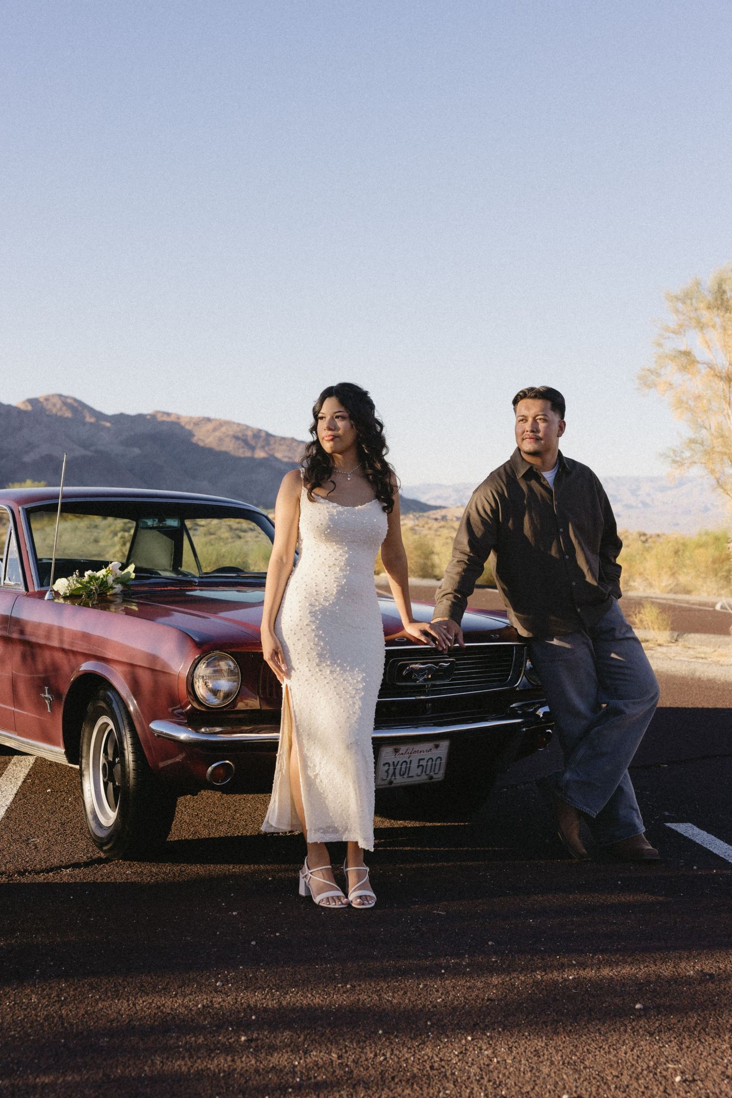 Couple poses with vintage burgundy car in desert landscape.