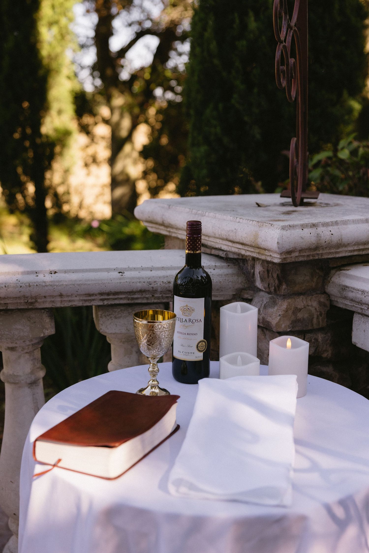 Outdoor wedding ceremony table with red wine bottle, gold chalice, Bible, white candles on white cloth with stone balustrade backdrop.