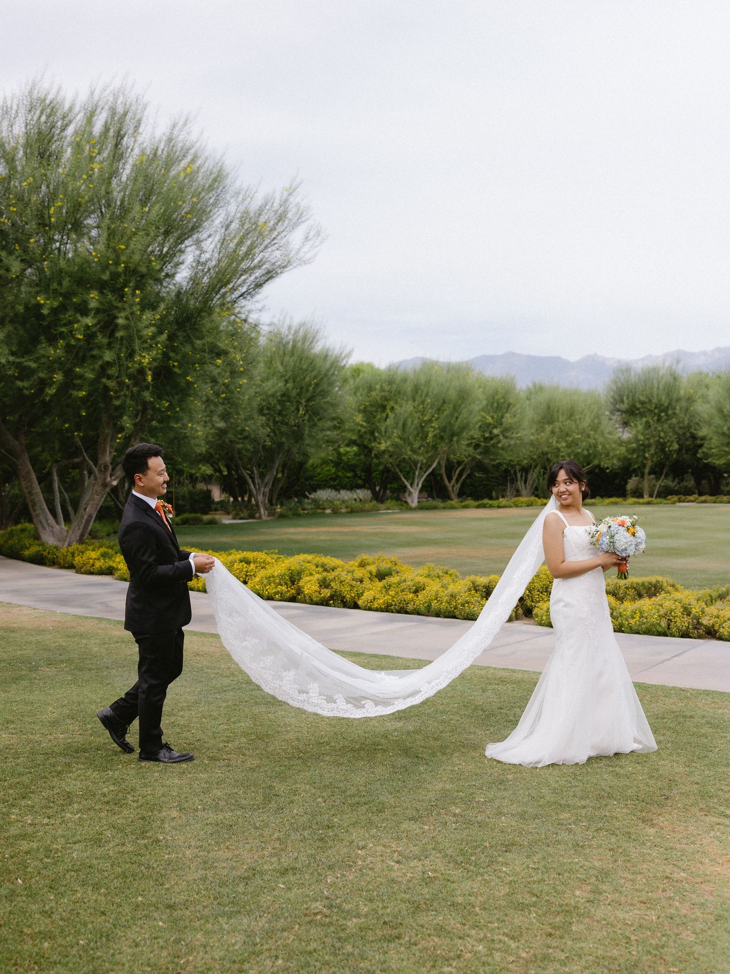 Bride and groom pose on lush green lawn, her long veil trailing behind them, with mountain views and trees.