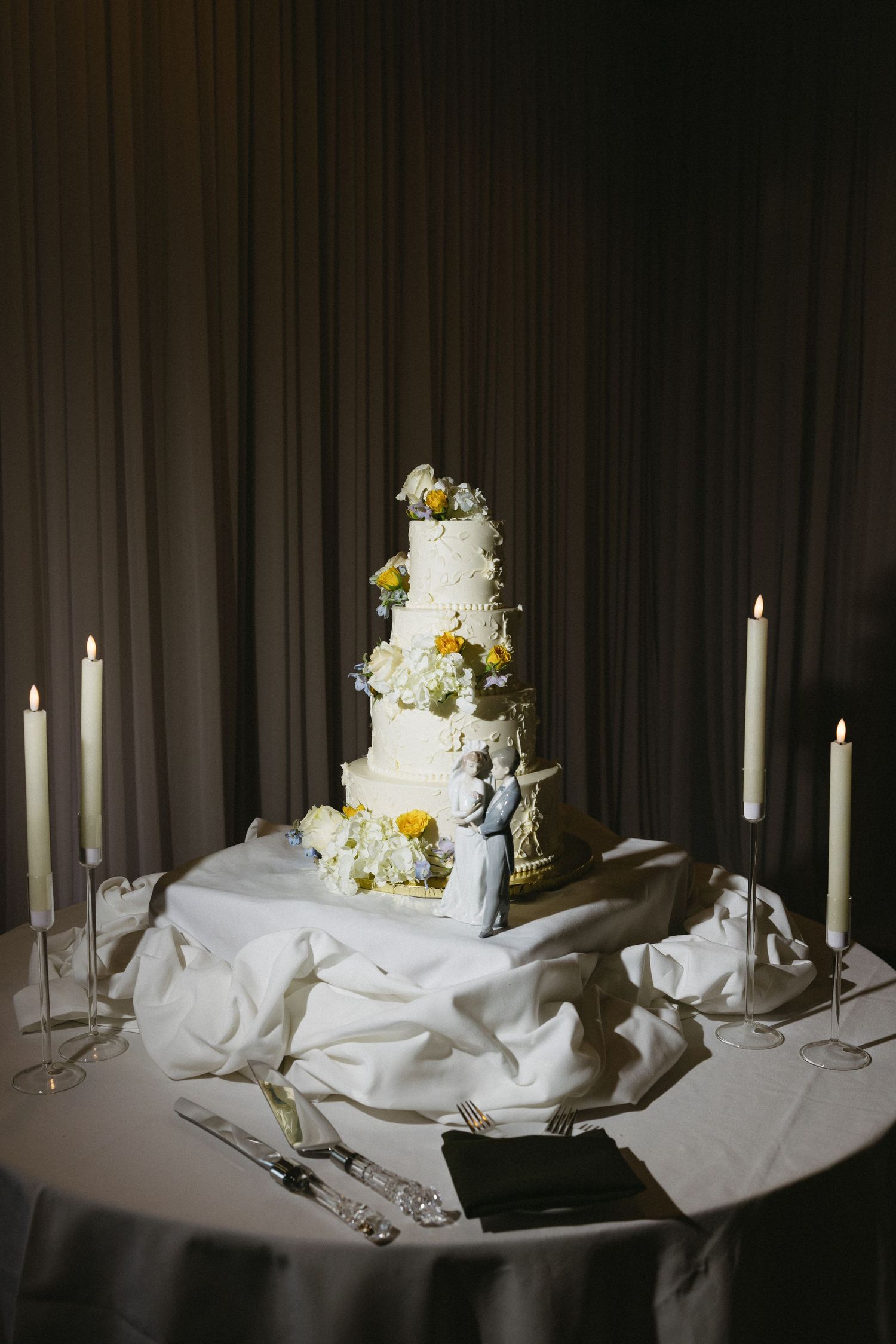 Elegant four-tier white wedding cake with floral decor and bride-groom topper, surrounded by tall candles on draped table.