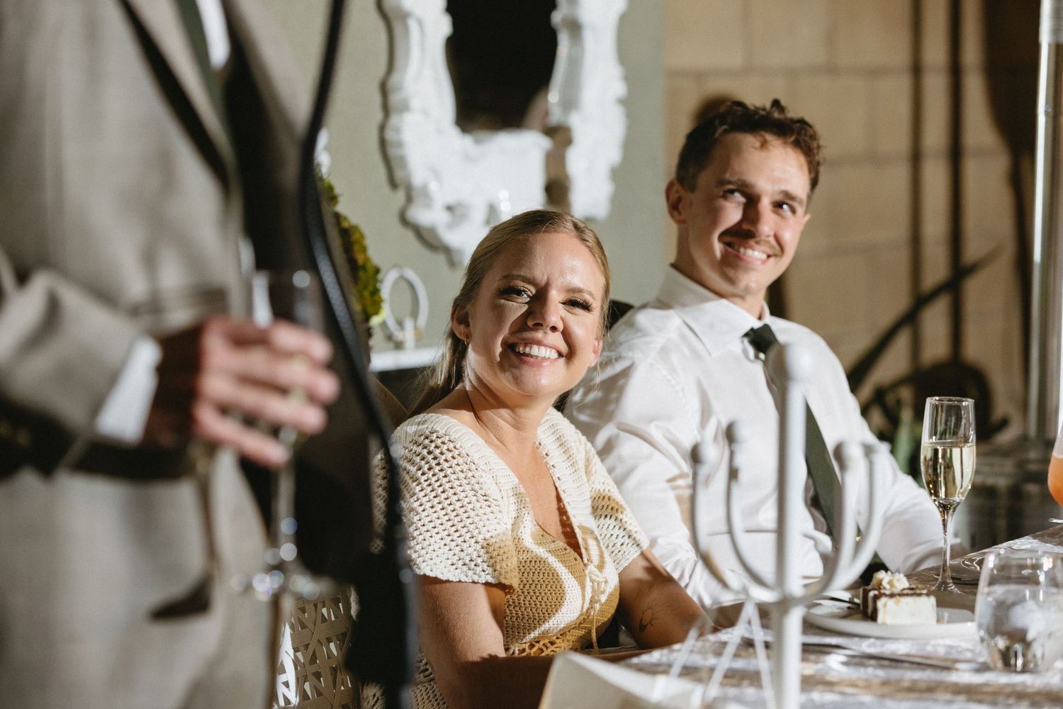 Smiling couple seated at a wedding reception table, woman in crochet top and man in white shirt with bow tie.