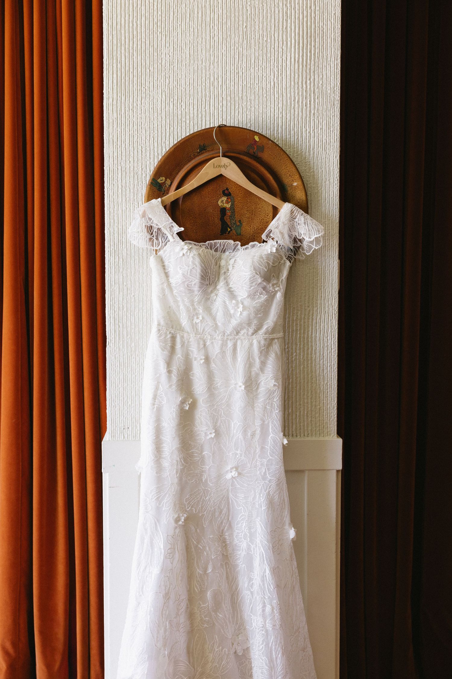 White lace off-shoulder wedding dress hanging on wooden hanger against textured wall with orange curtains.