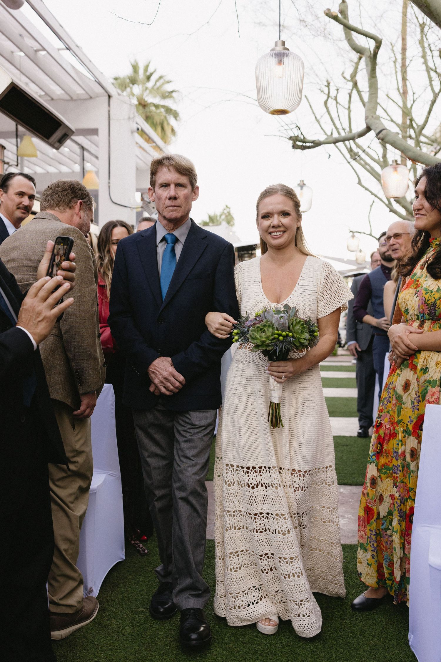 Bride in lace dress walks down outdoor aisle with escort, holding bouquet, guests watching at garden wedding ceremony.