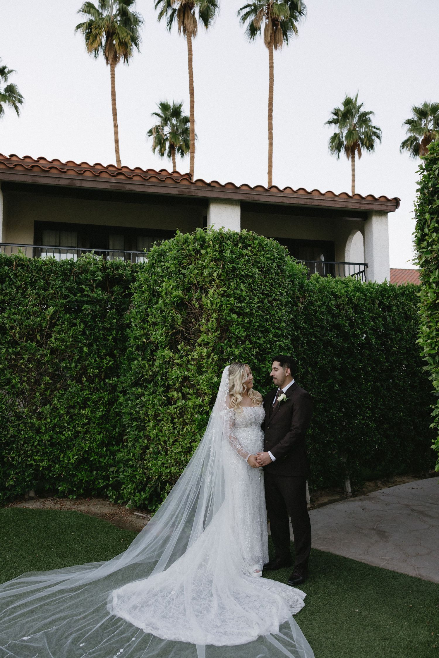 Wedding couple posing in garden setting with palm trees and flowing veil.