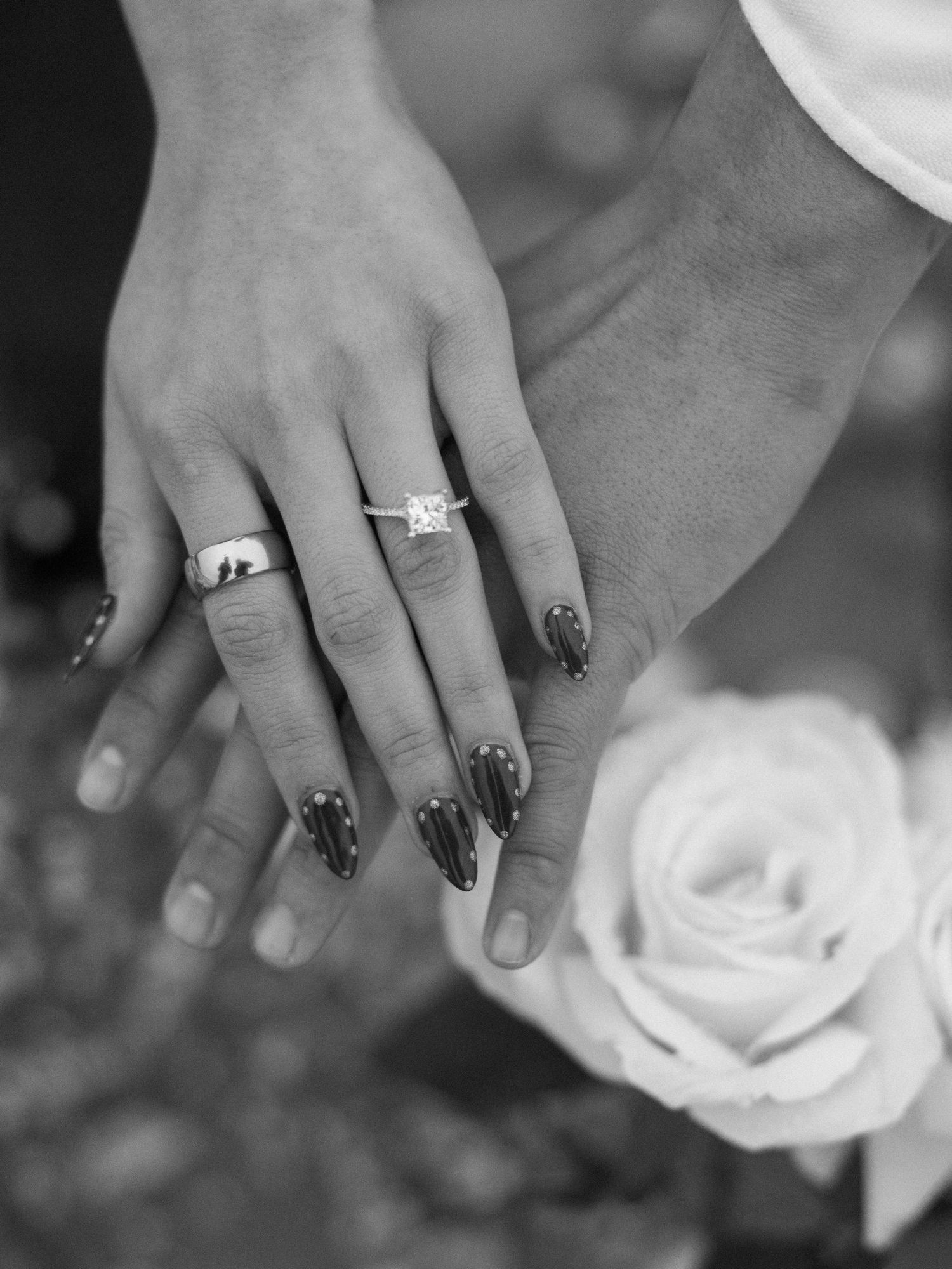 Close-up of hands with engagement ring and black nail polish next to white rose.