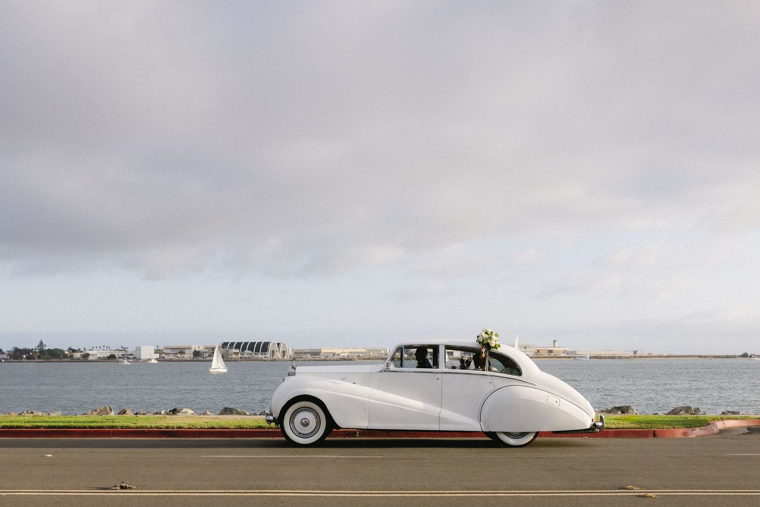 Classic white vintage car parked near the ocean with cloudy skies in the background.