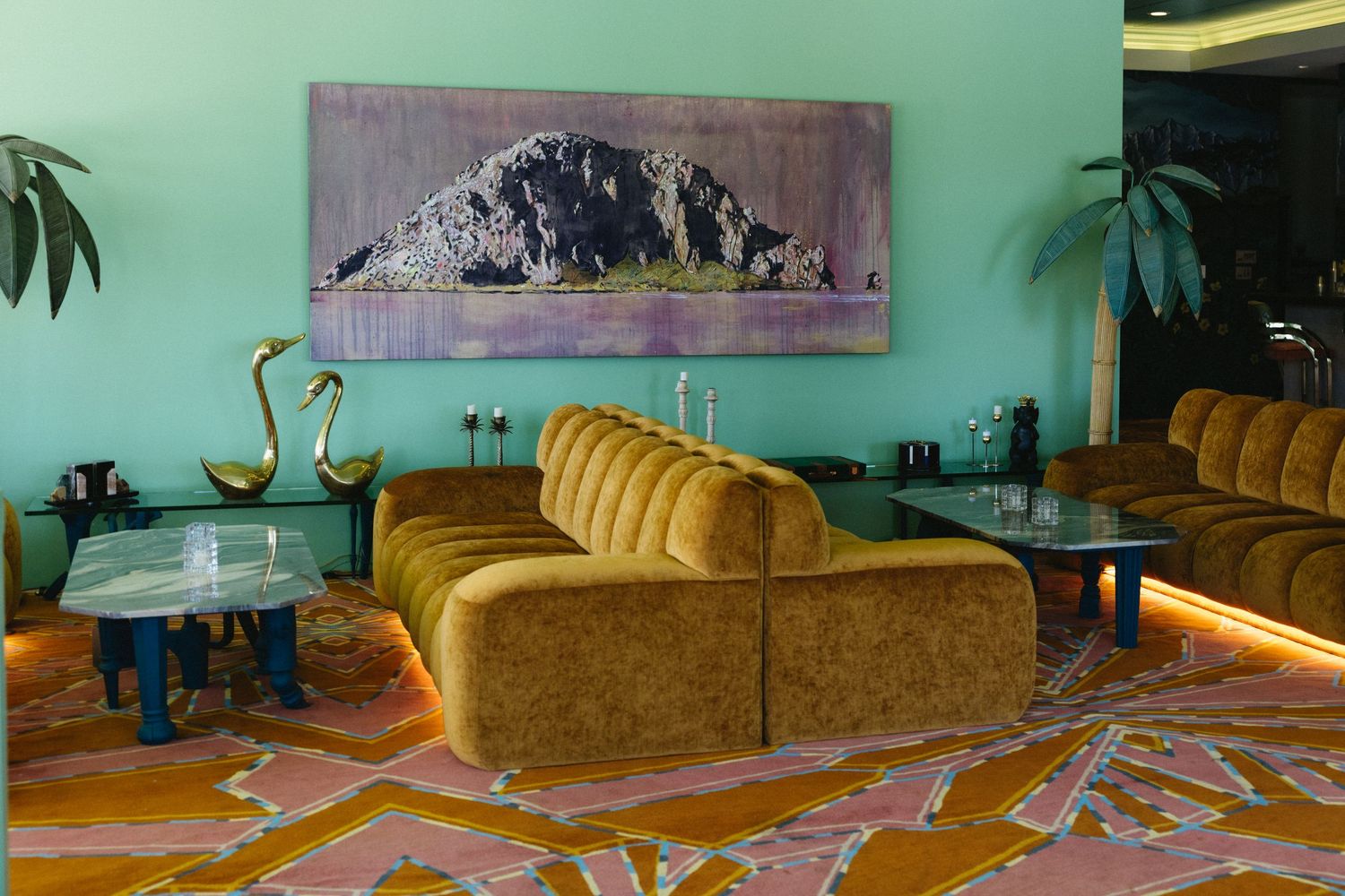 Vibrant hotel lounge with mustard velvet sofas, teal walls, colorful patterned carpet, and a mountain landscape painting.