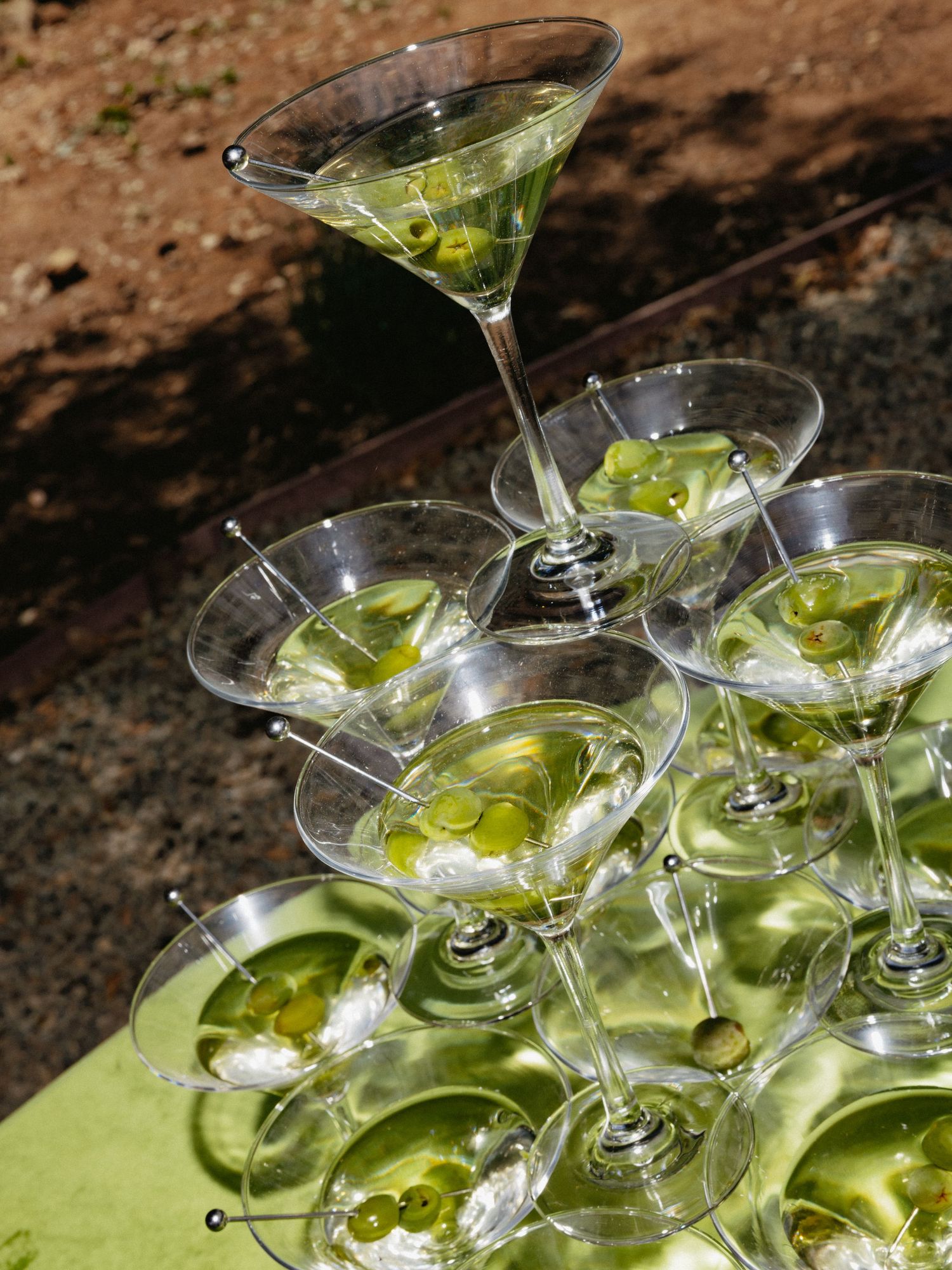 A pyramid tower of martini glasses filled with green olive garnished cocktails displayed on a green tablecloth outdoors.