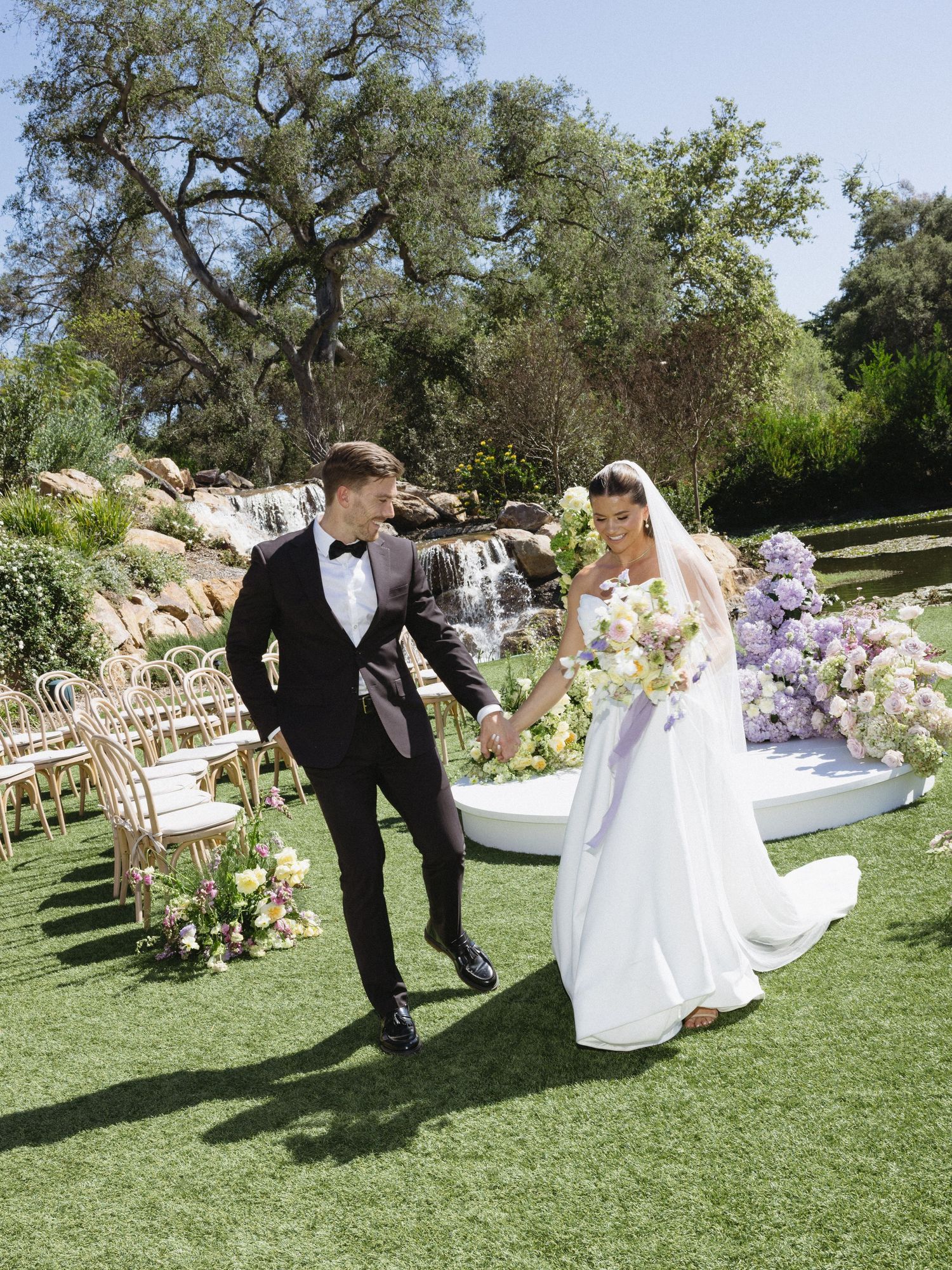 Bride and groom walk hand in hand across green lawn after outdoor ceremony with floral arch and waterfall backdrop.