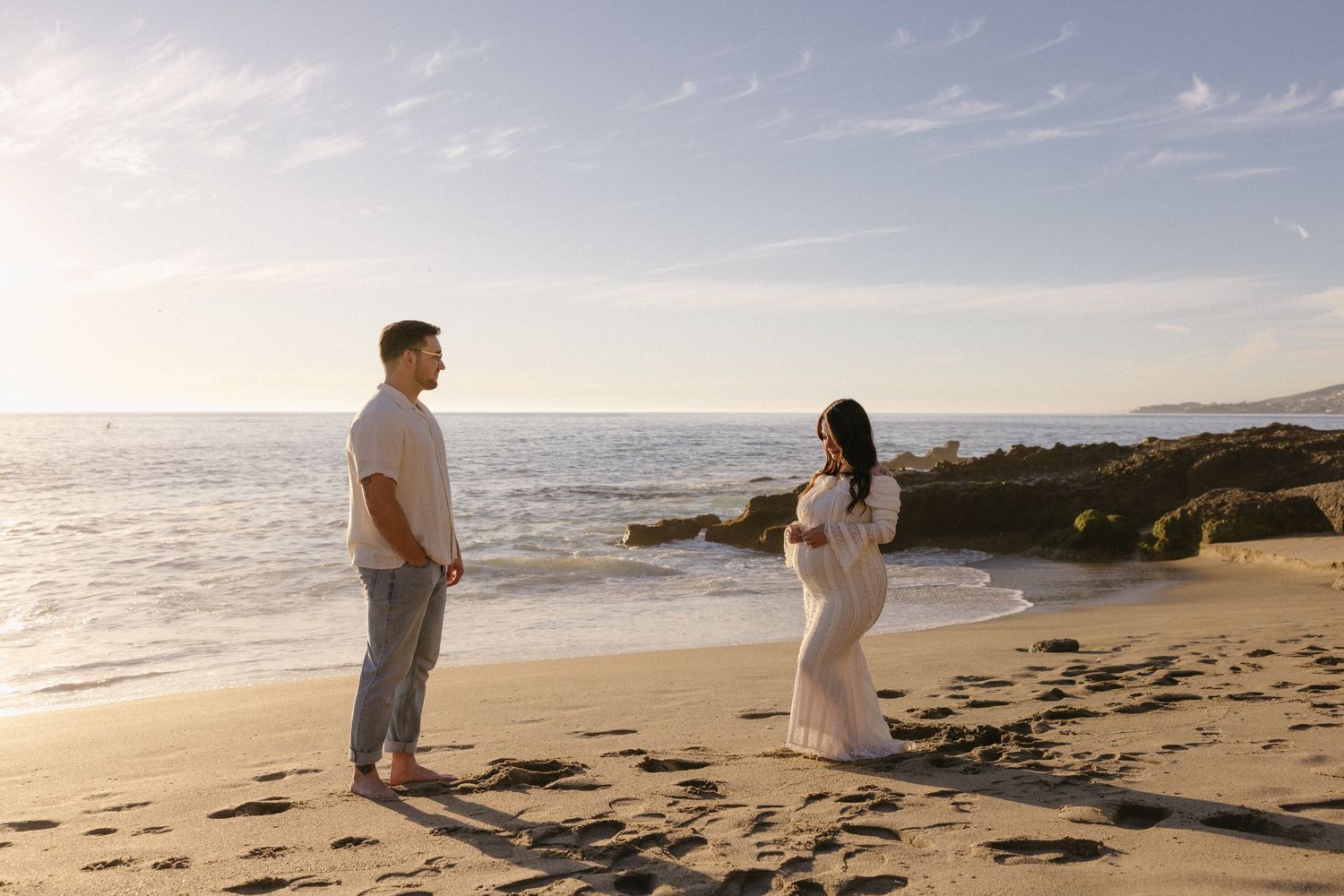 A couple strolls along a sandy beach with waves and cliffs in the distance.