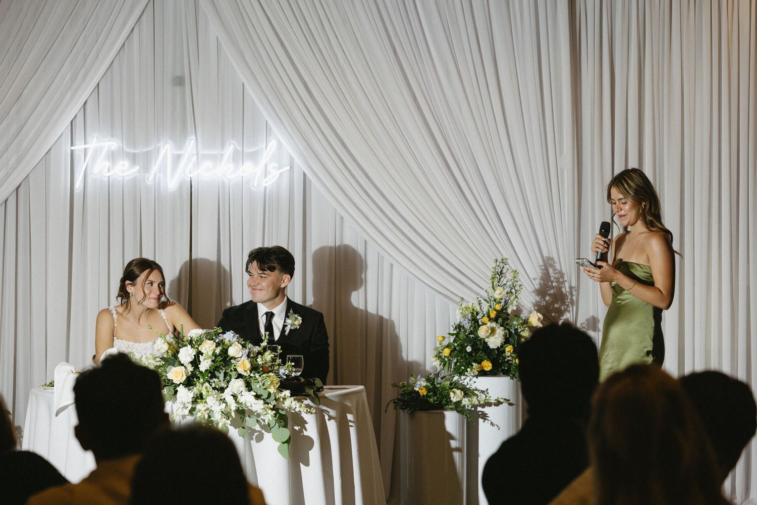 Wedding reception speech with bride and groom seated at floral table, bridesmaid speaking at microphone under neon sign.