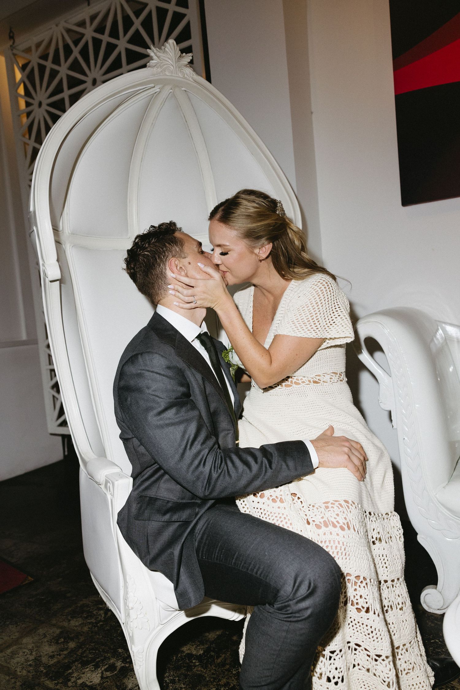 A bride and groom share a romantic kiss while seated on an elegant white throne chair at their wedding reception.