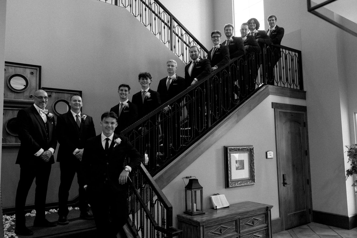 Groomsmen pose on elegant staircase in black and white wedding photo inside a formal venue.