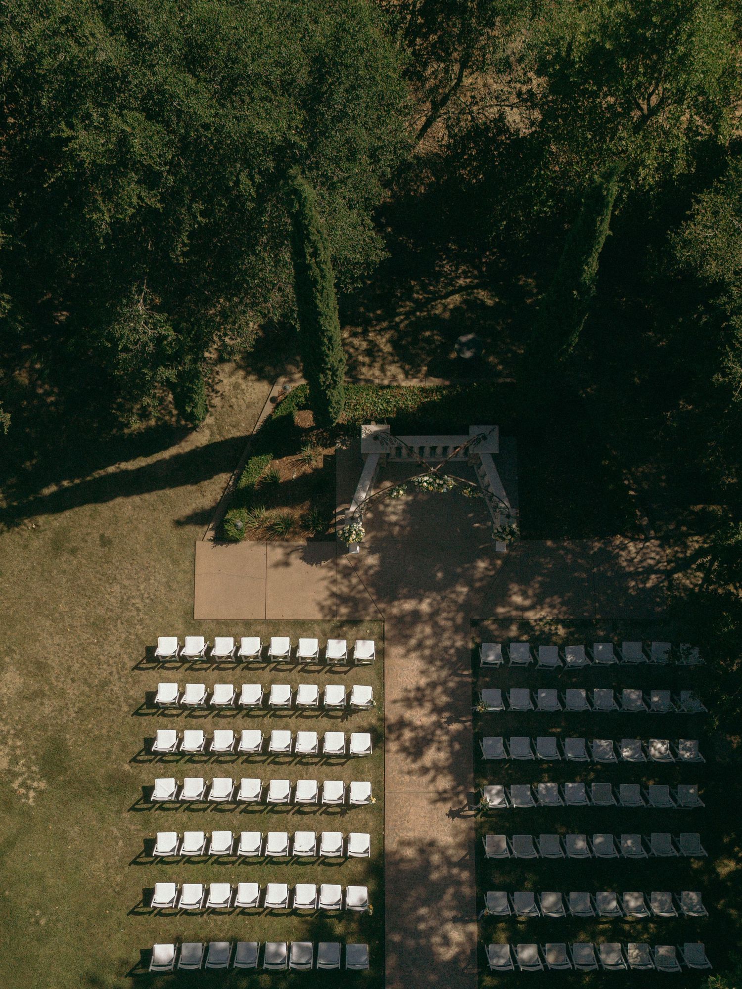 Aerial view of an outdoor wedding ceremony setup with rows of white chairs and a floral arch altar surrounded by lush trees.