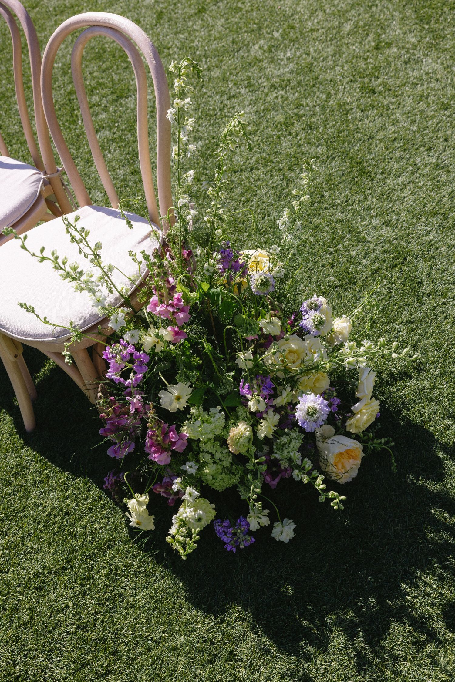 Wildflower wedding aisle arrangement with purple and white blooms beside bentwood chairs on grass.
