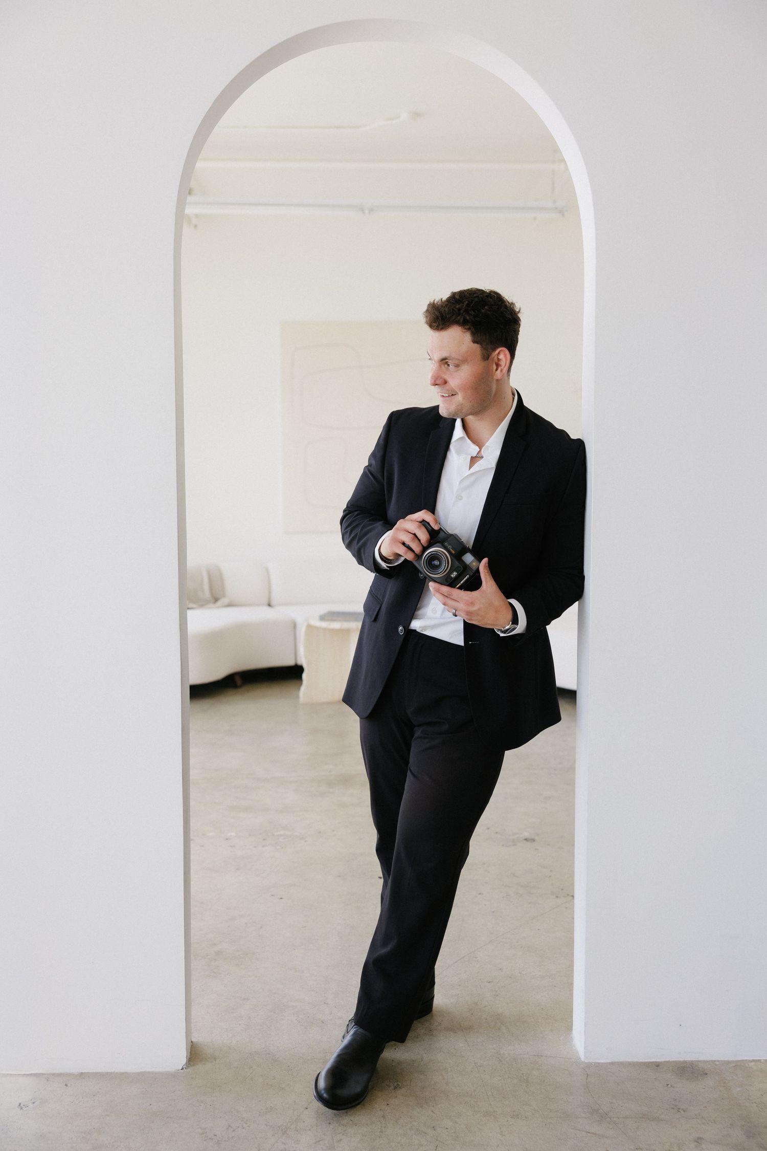 A man in a black suit holds a camera while standing in a white arched doorway, smiling confidently.