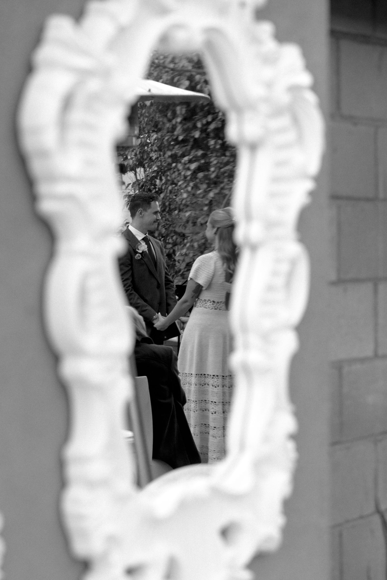 Black and white wedding photo of bride and groom holding hands, reflected in an ornate vintage mirror outdoors.