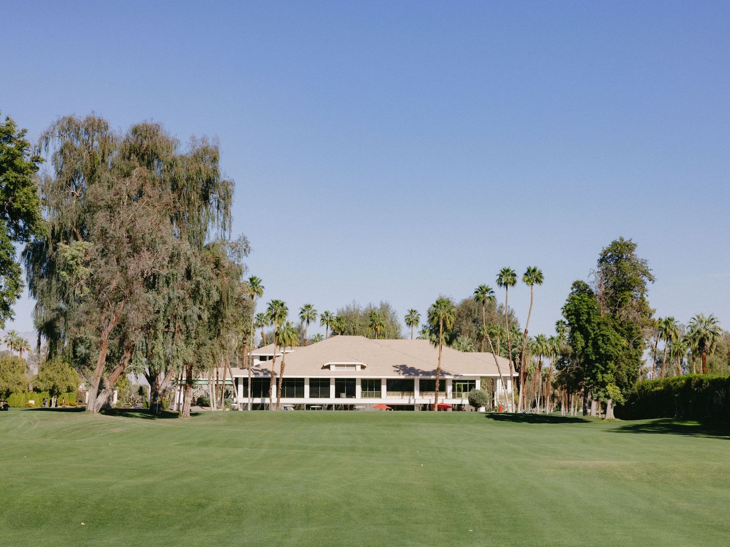 Lush green golf course with a sprawling clubhouse surrounded by mature trees under a clear blue sky.