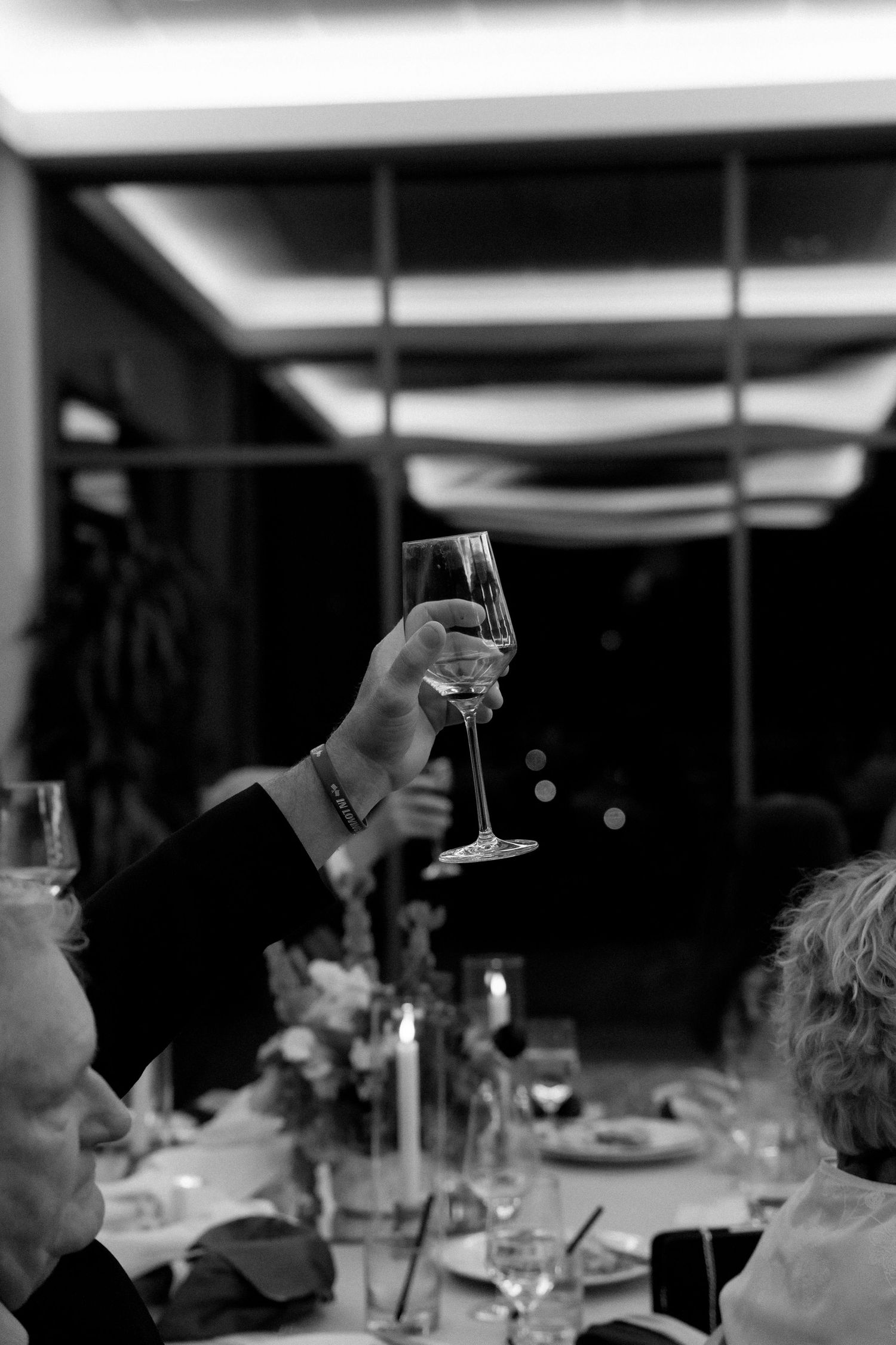 Black and white photo of a hand raising a champagne flute at an elegant dinner party with candles and floral centerpieces.