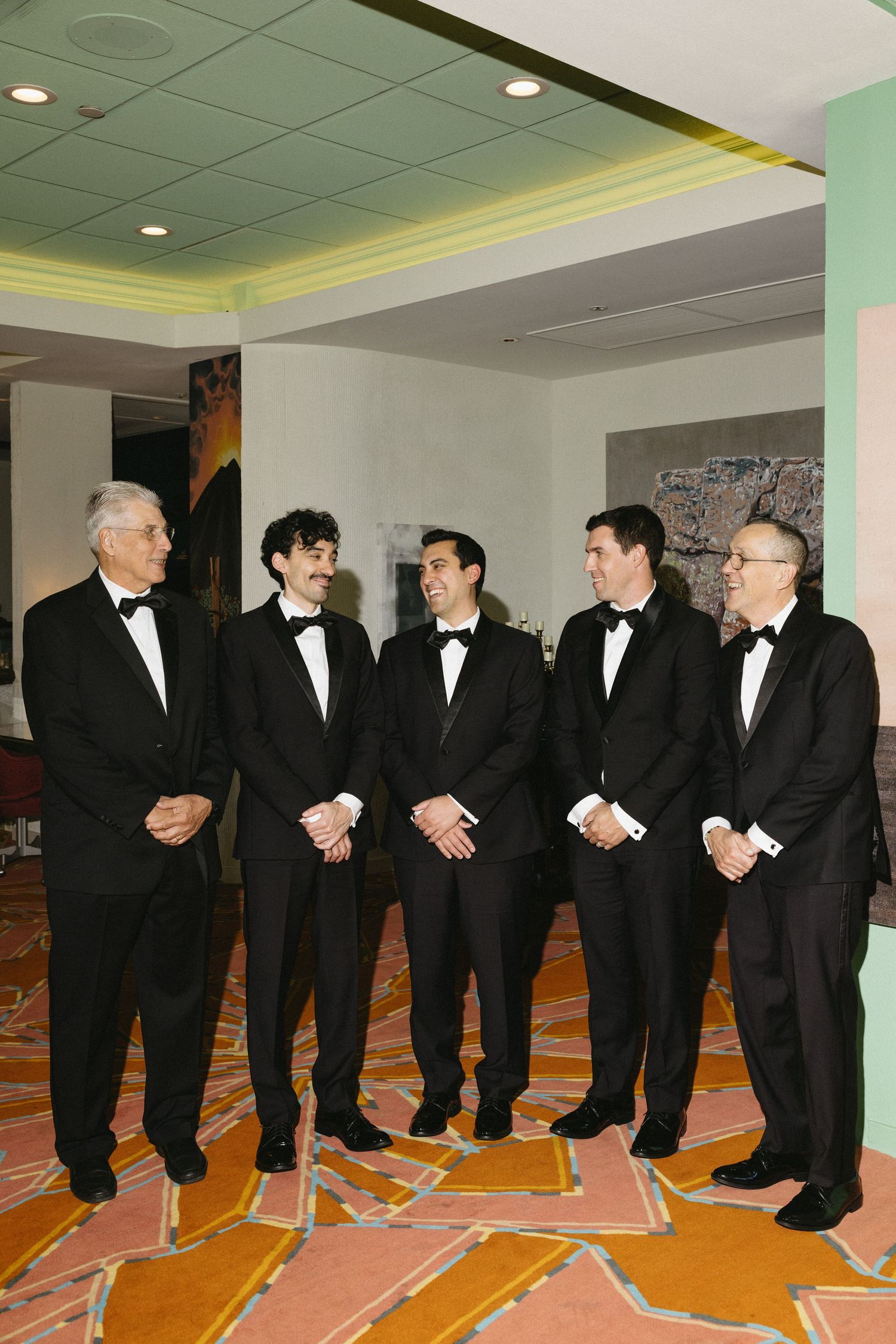 Five groomsmen in black tuxedos smiling together in elegant hotel lobby interior.