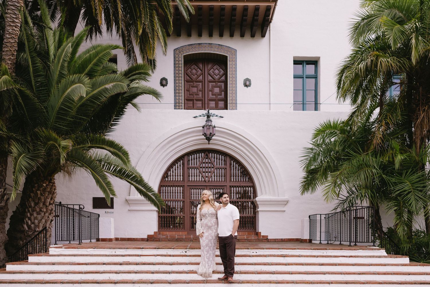 White Spanish colonial architecture with arched doorways and palm trees line a courtyard.