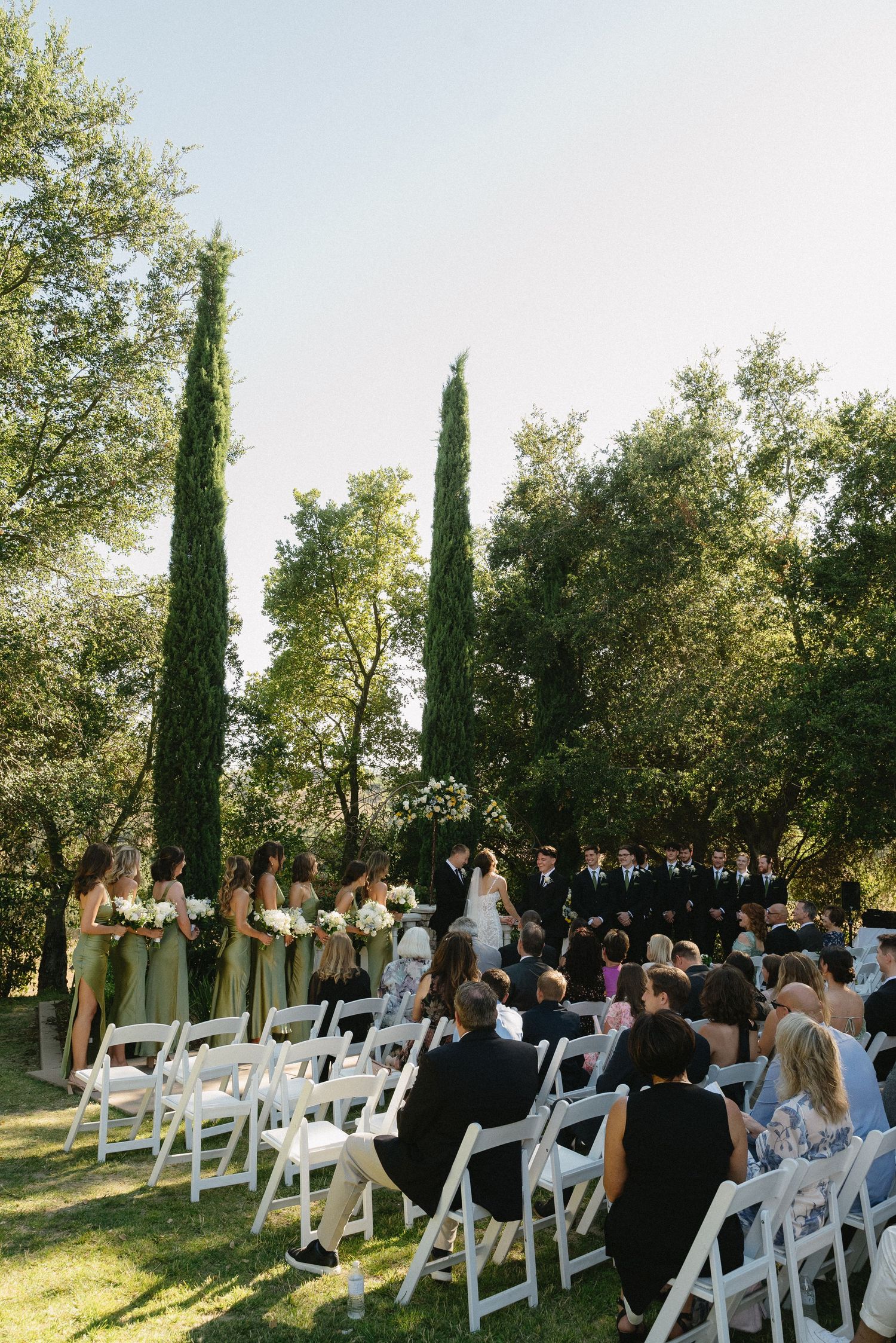 Outdoor wedding ceremony with guests seated on white chairs, bridal party at altar, surrounded by tall cypress and lush green trees.