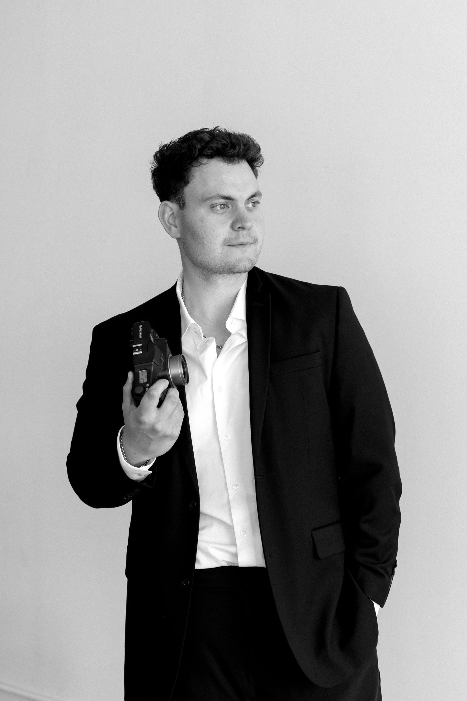 Black and white portrait of a young man in a suit holding a camera, looking confidently to the side.