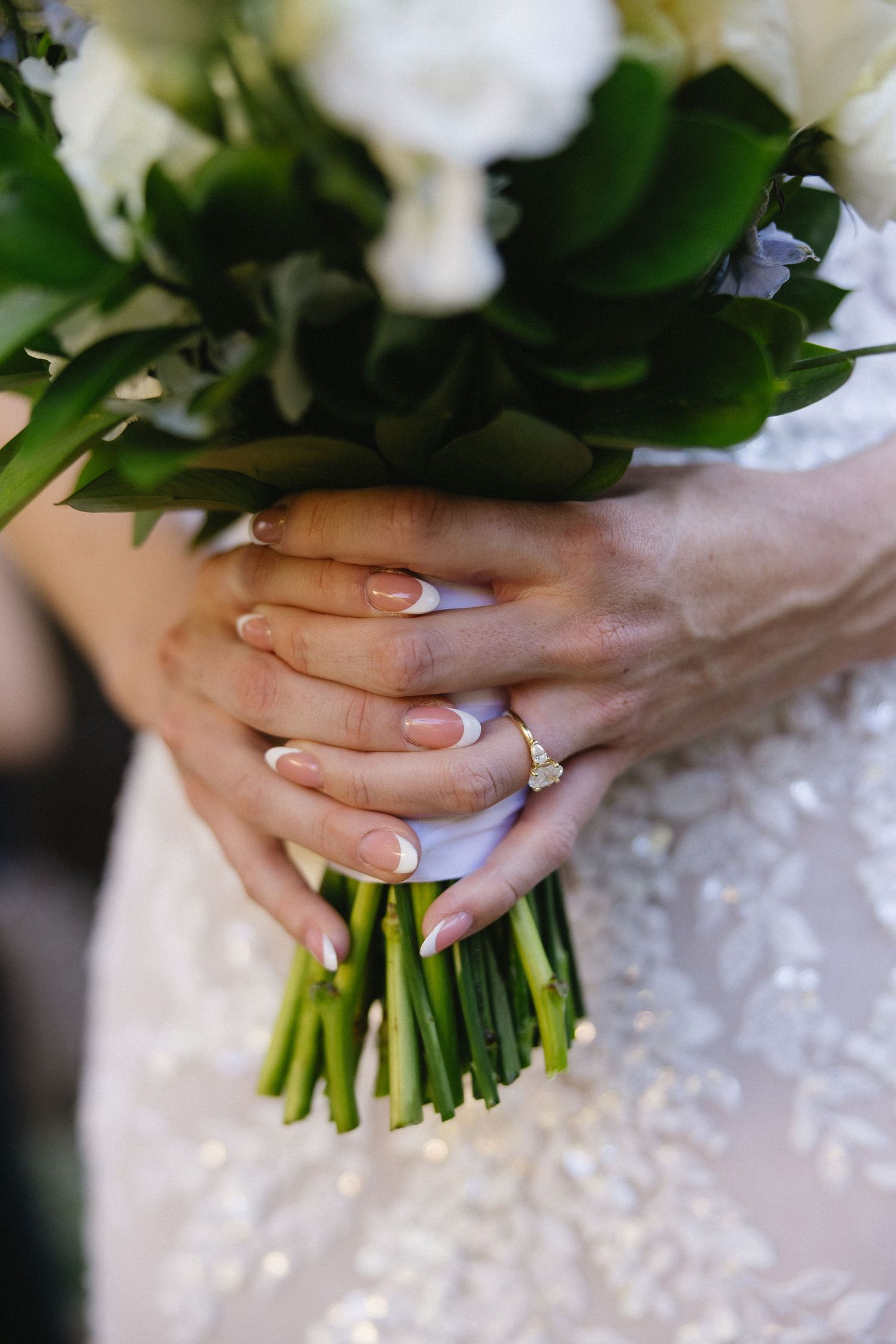 Bride holding white floral wedding bouquet with french manicured nails and diamond engagement ring on ring finger.