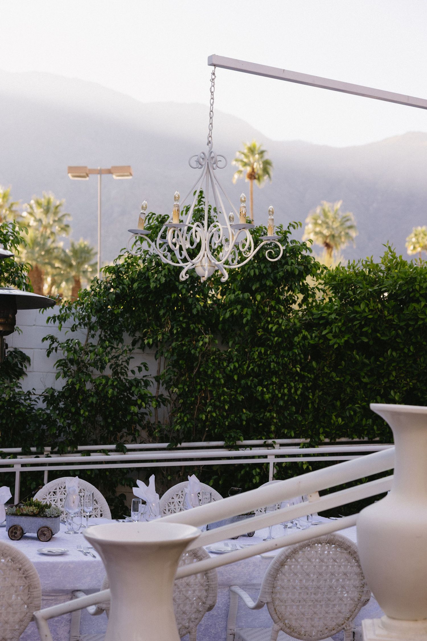 Elegant outdoor wedding reception with white chandelier, wicker chairs, and palm trees against mountain backdrop.