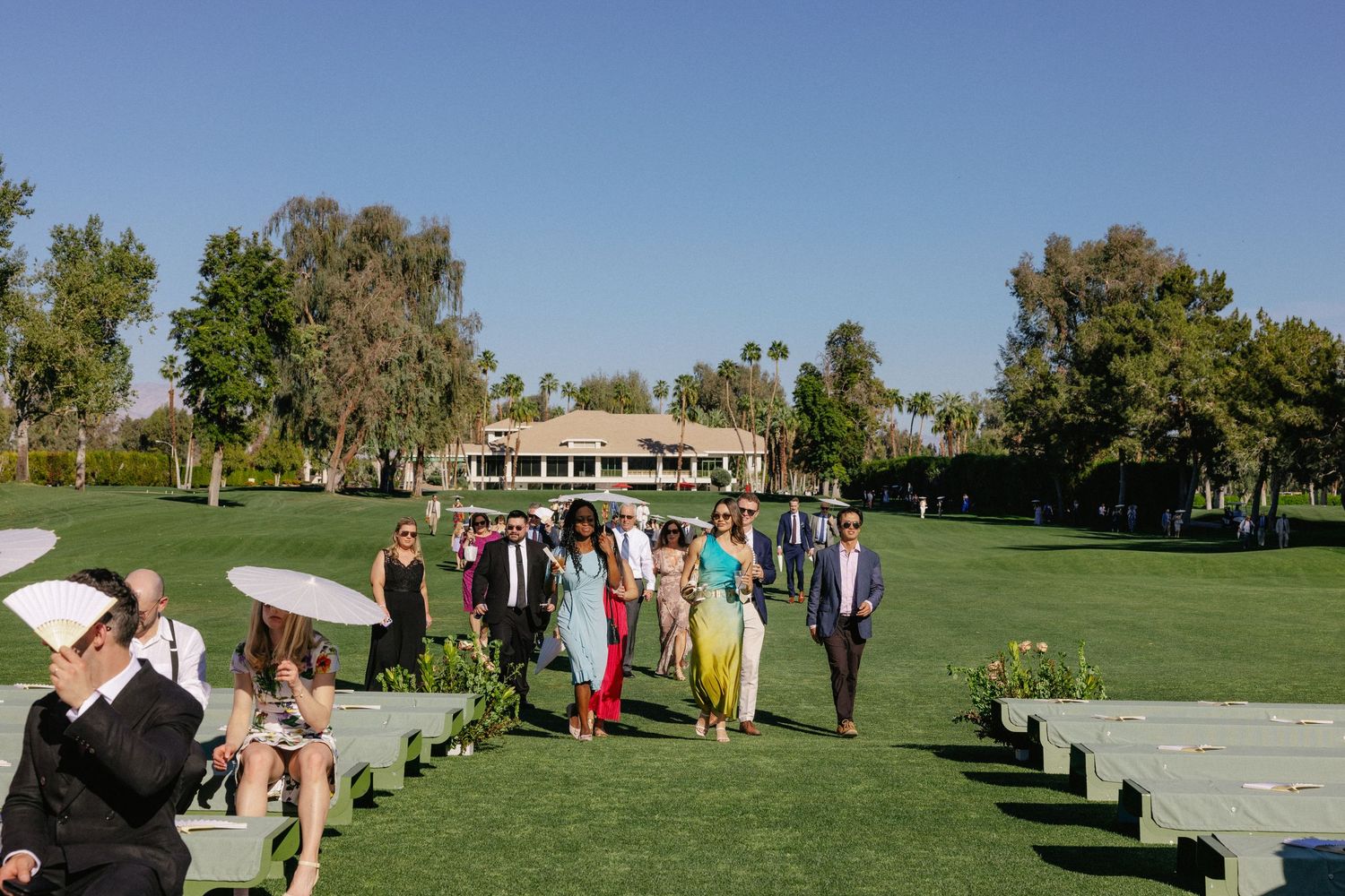 Wedding guests walk across lush green lawn at outdoor ceremony venue surrounded by palm trees on sunny day.