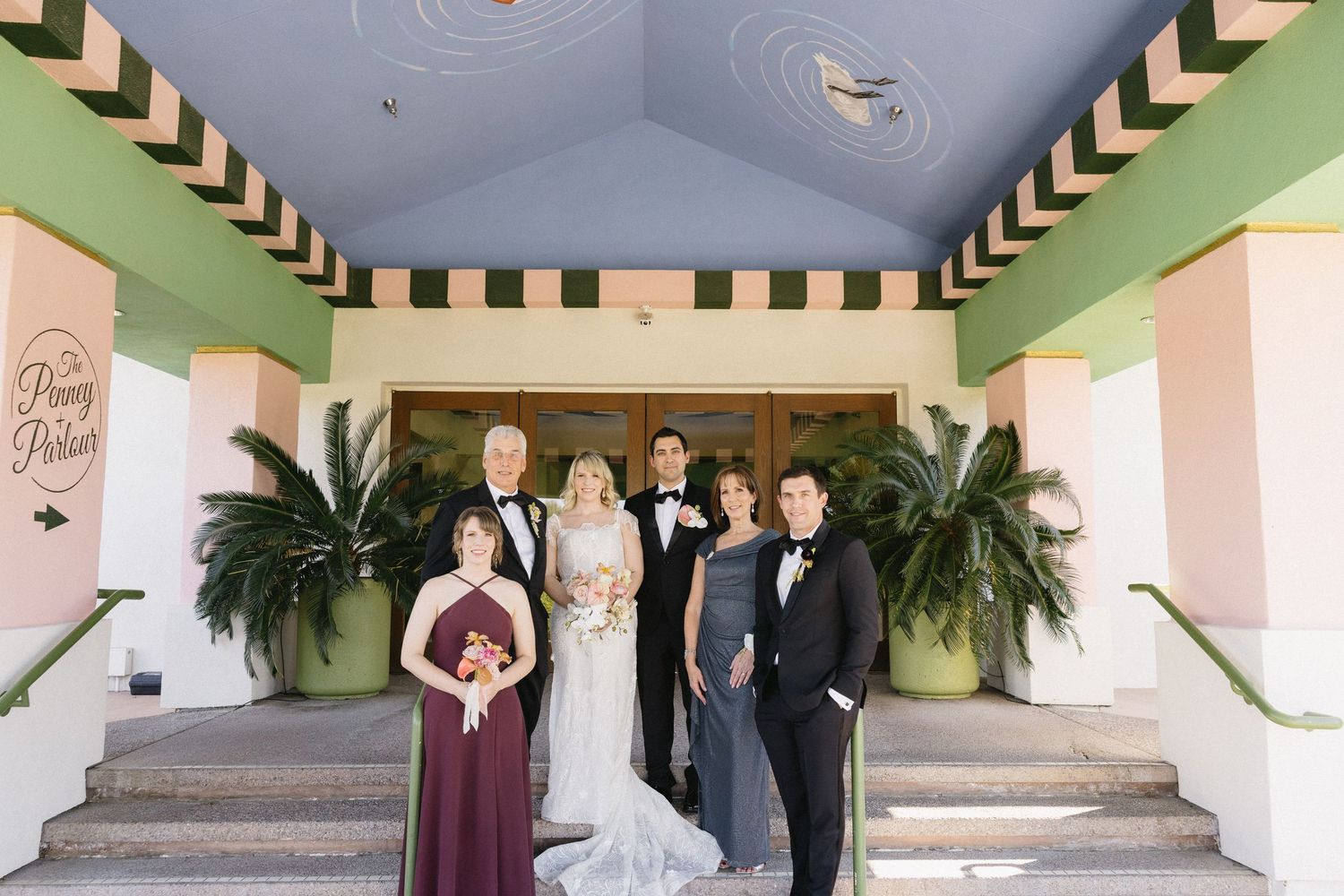 Wedding party poses on steps outside a colorful venue with pink and green walls and a blue ceiling.