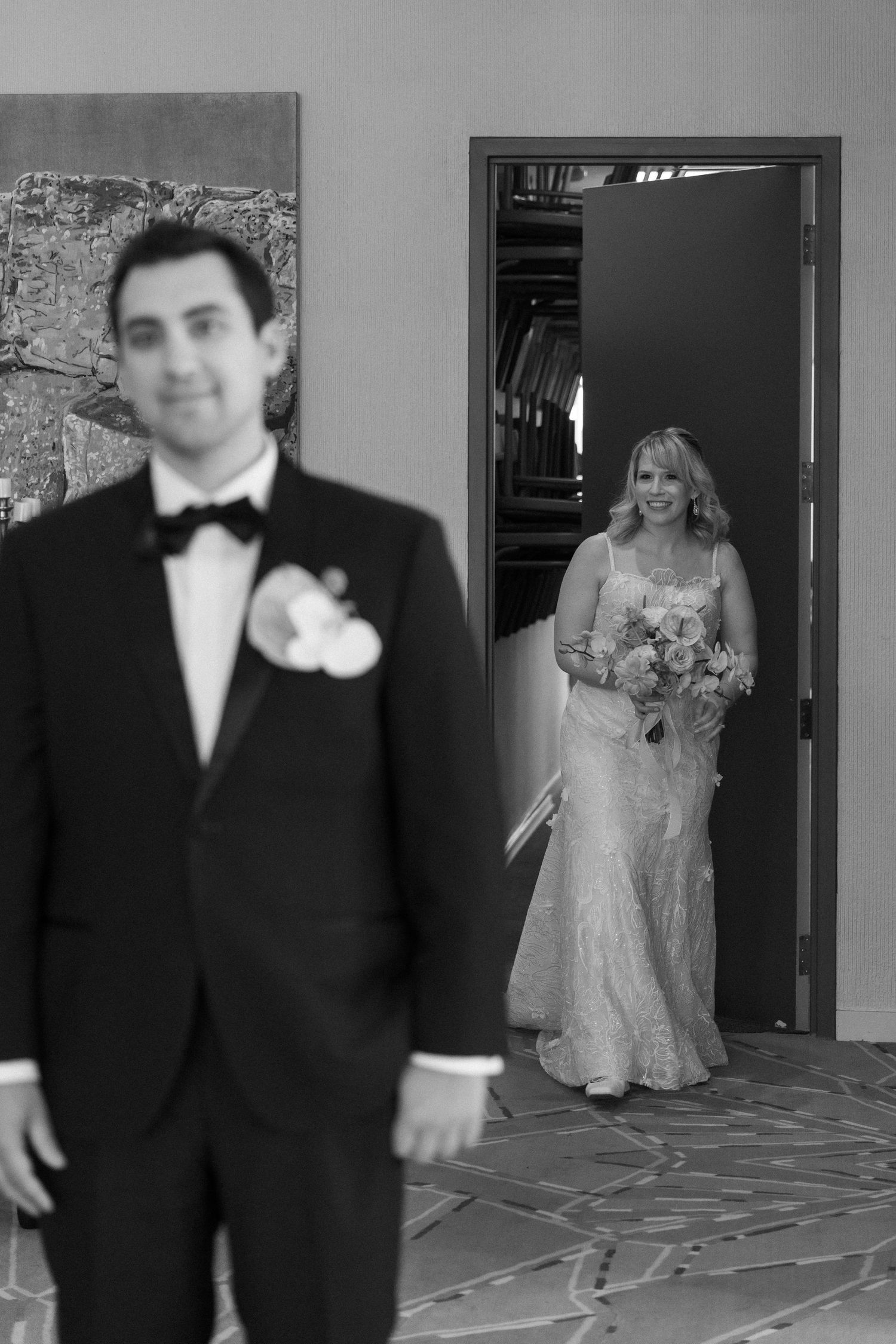 Black and white wedding photo of groom in tuxedo with bride in lace gown holding bouquet approaching through doorway.