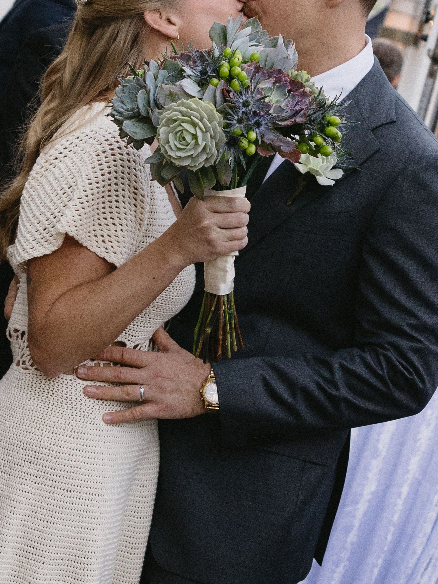 Bride and groom kissing while holding a succulent and berry wedding bouquet, bride wearing a white crochet dress.