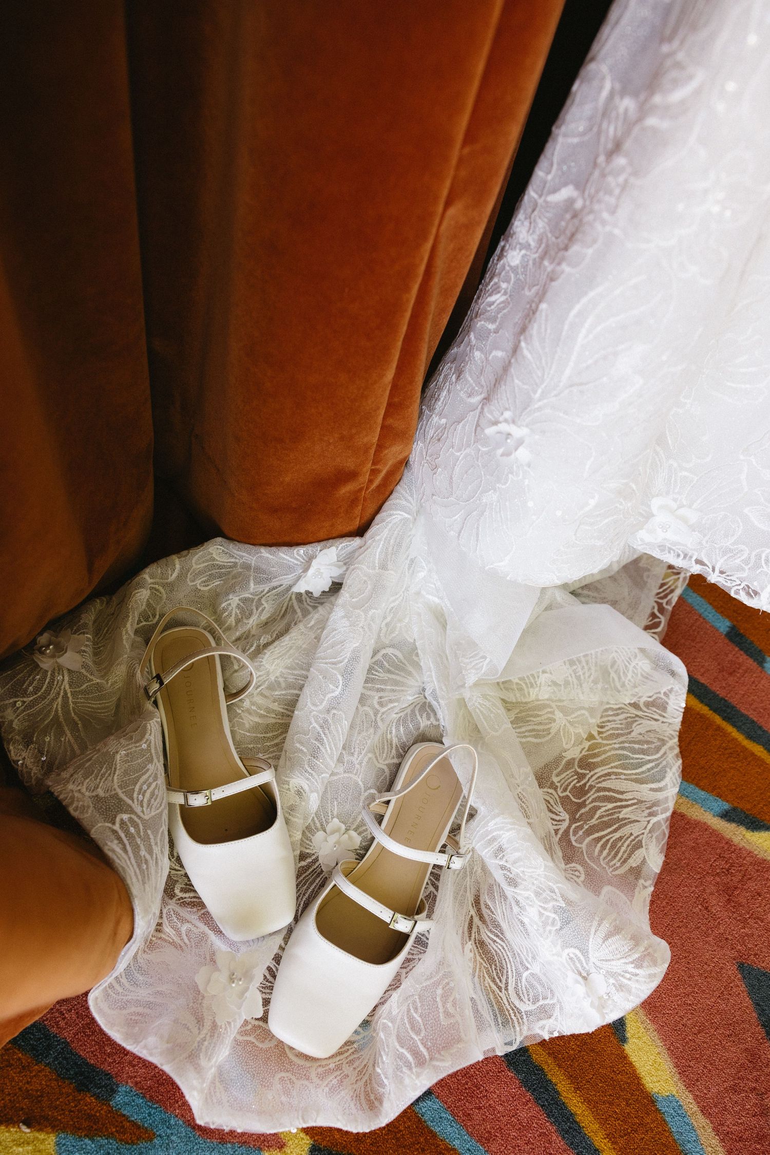 White bridal heels with ankle straps rest on lace wedding dress fabric beside an orange velvet chair.