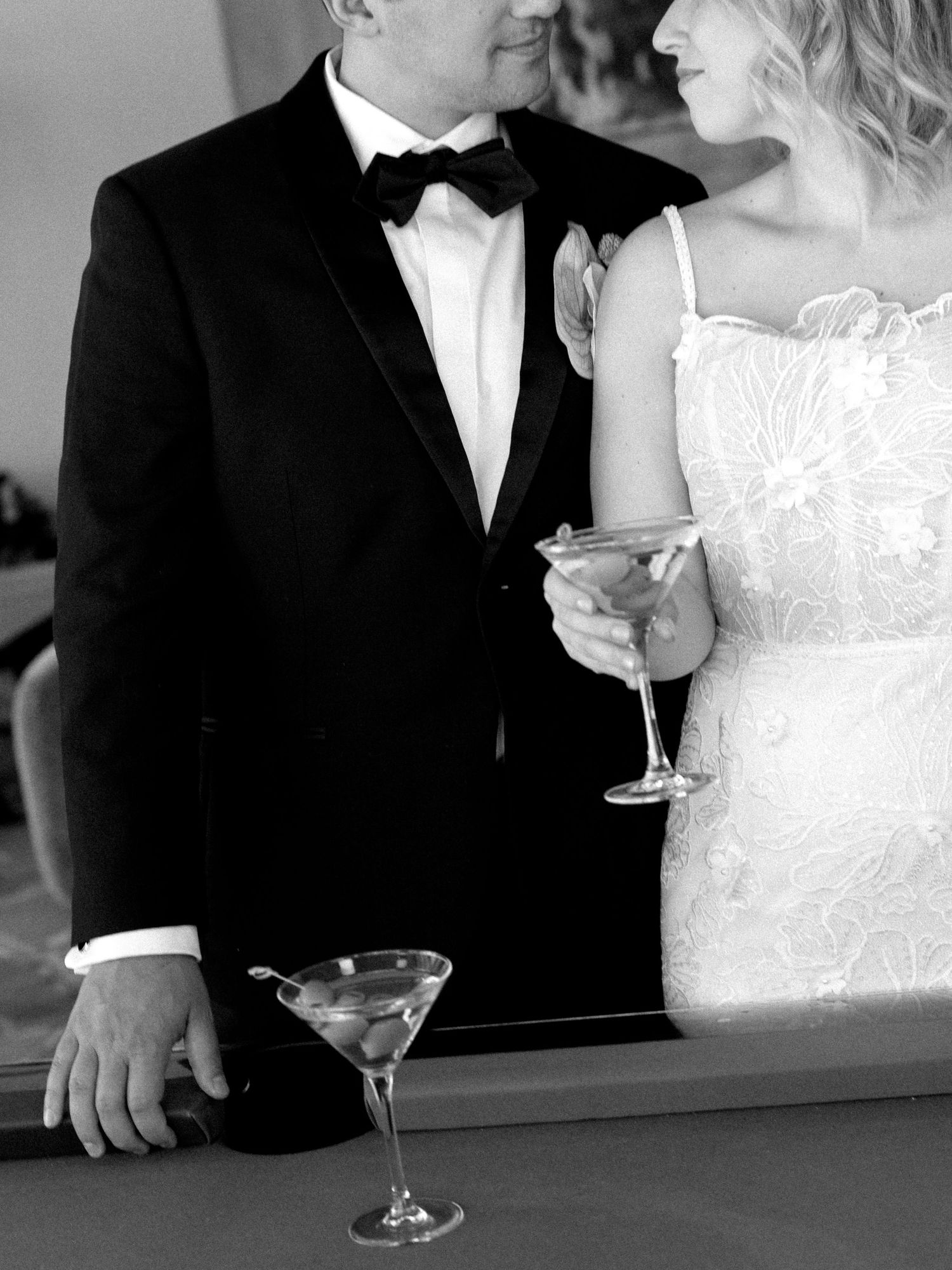 Black and white wedding photo of bride and groom holding martini glasses at a bar in elegant formal attire.