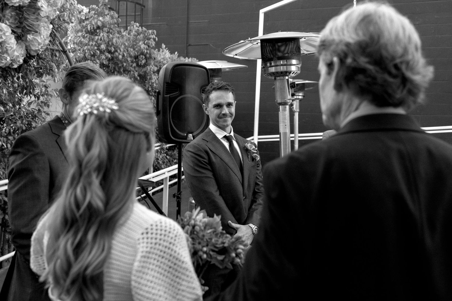 Black and white wedding ceremony photo showing groom smiling at bride as officiant presides outdoors.