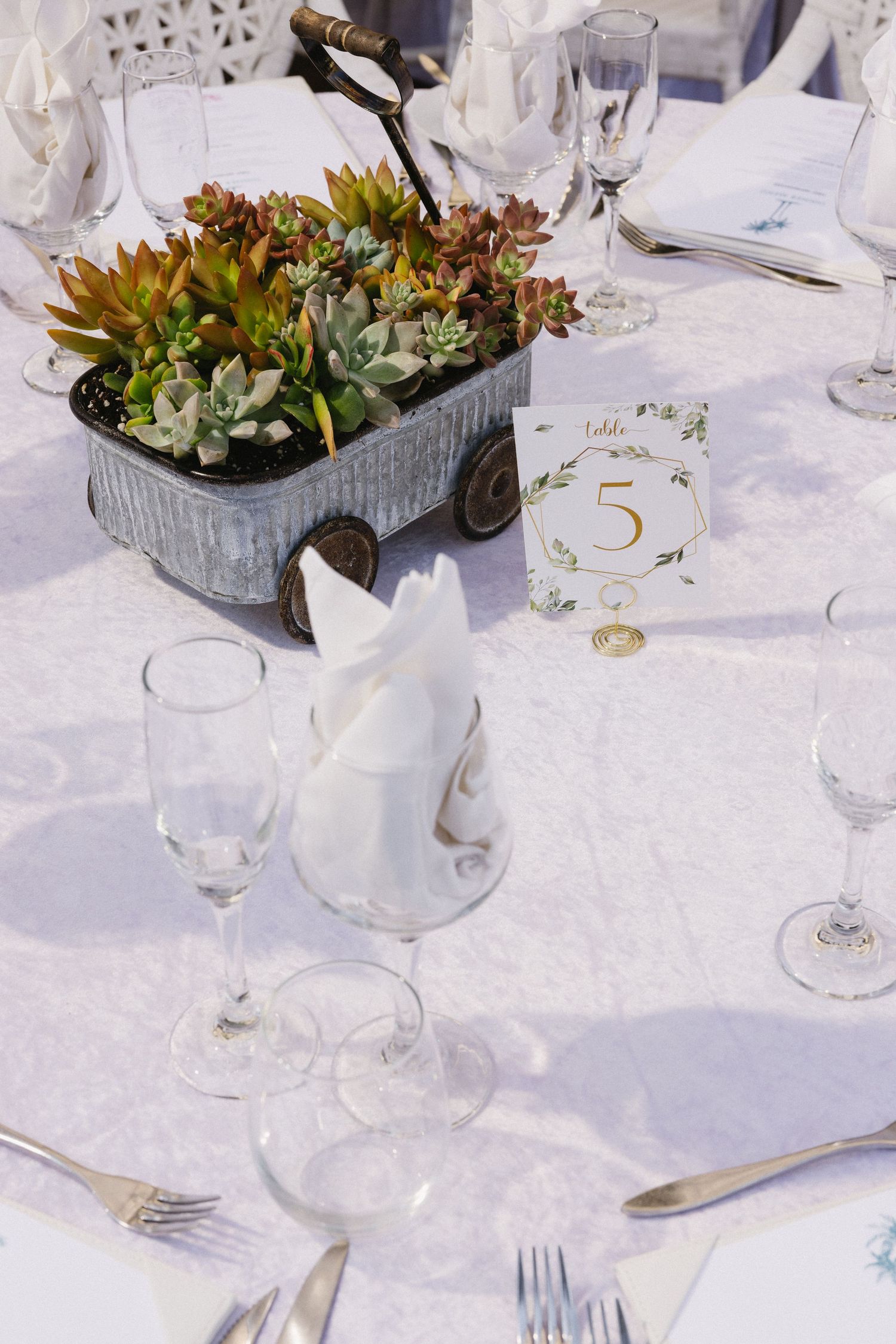 Elegant wedding reception table five with succulent centerpiece in vintage wagon, white linens, crystal glasses, and folded napkins.