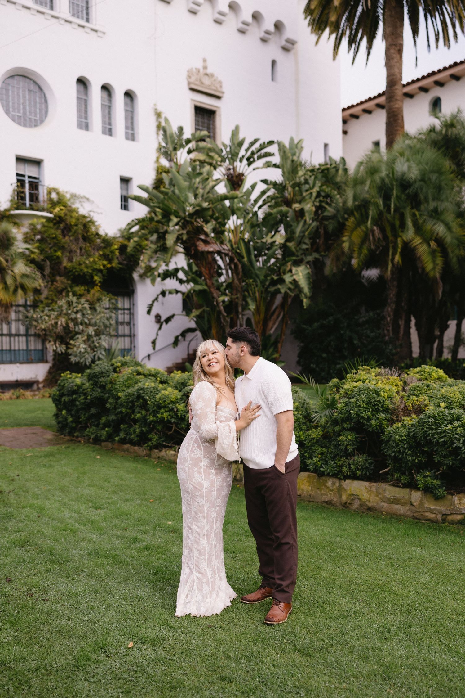 A couple shares a romantic moment on a grassy lawn in front of a Spanish-style building with palm trees.